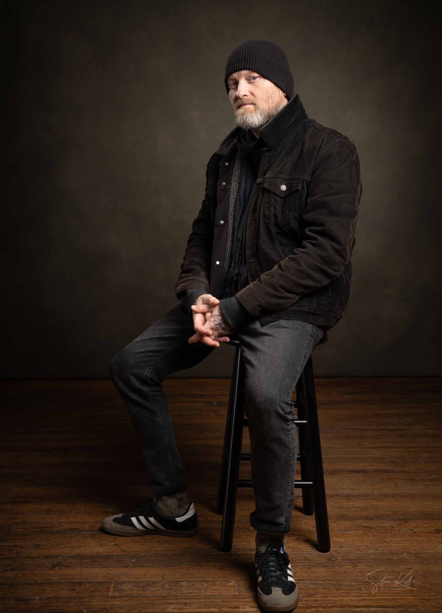 A figure in black winter clothing sits on a wooden chair in a dark studio setting with dramatic lighting.