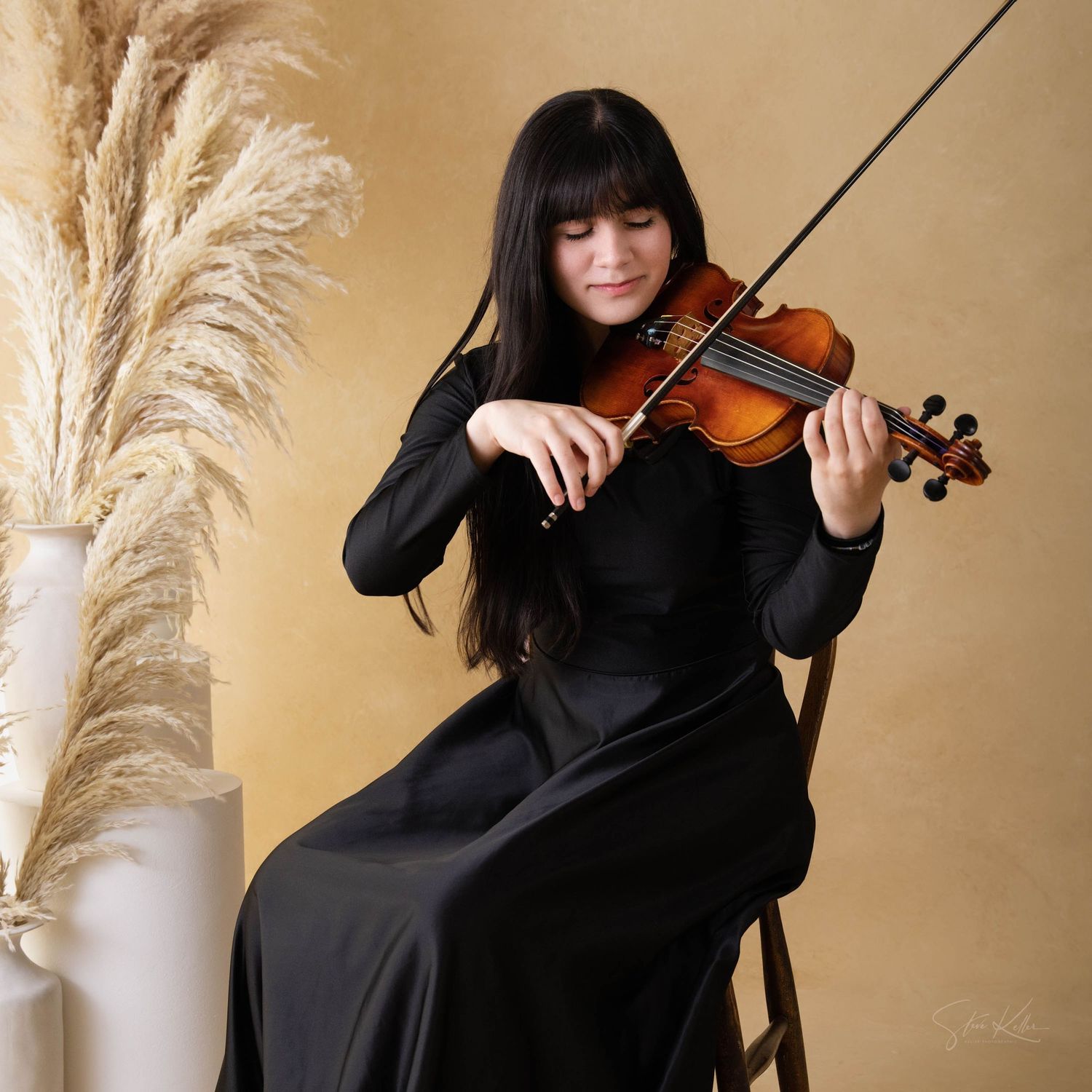 Musician in black dress plays violin while seated next to pampas grass against beige background.