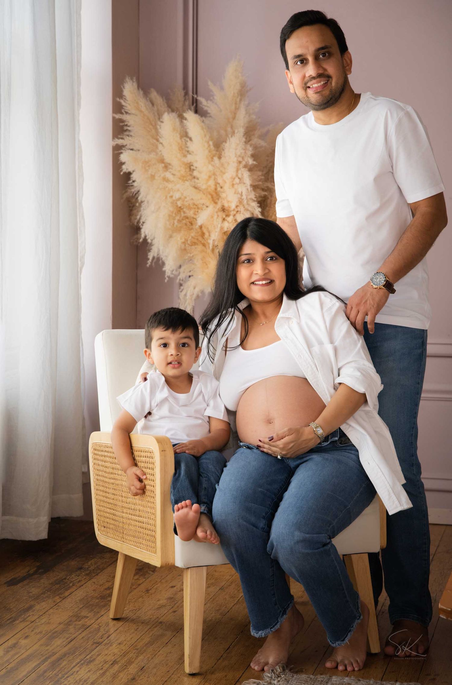 A pregnant woman sits in a chair with her toddler as her husband stands behind her for a maternity photo.