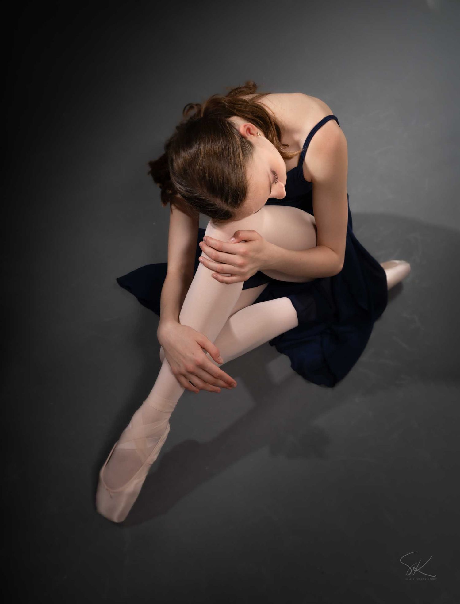 A ballerina in a navy dress sits gracefully on the floor hugging her knee in ballet pointe shoes.