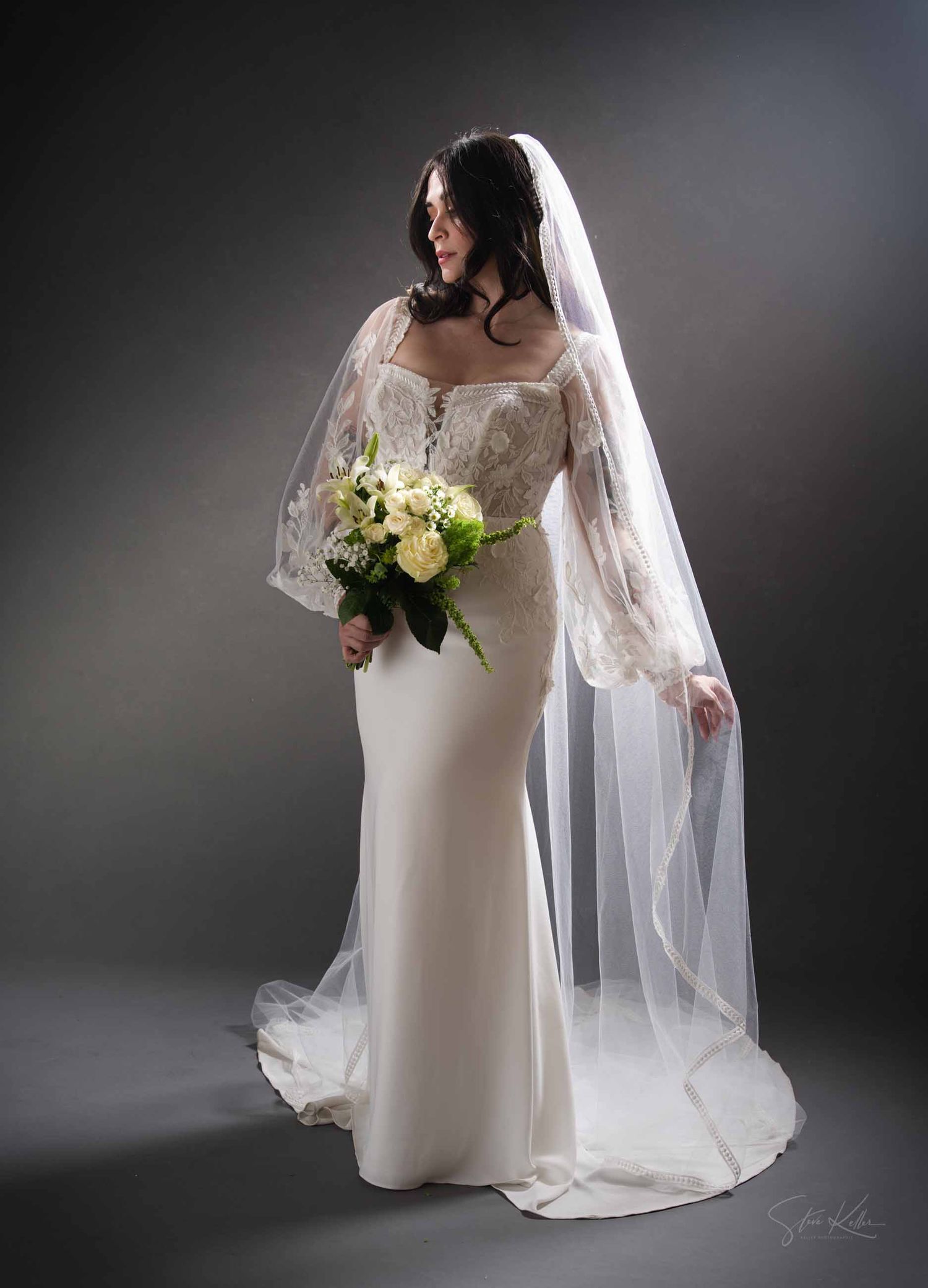 Elegant bride in flowing white gown and long veil poses gracefully against dark studio backdrop in artistic photo series.