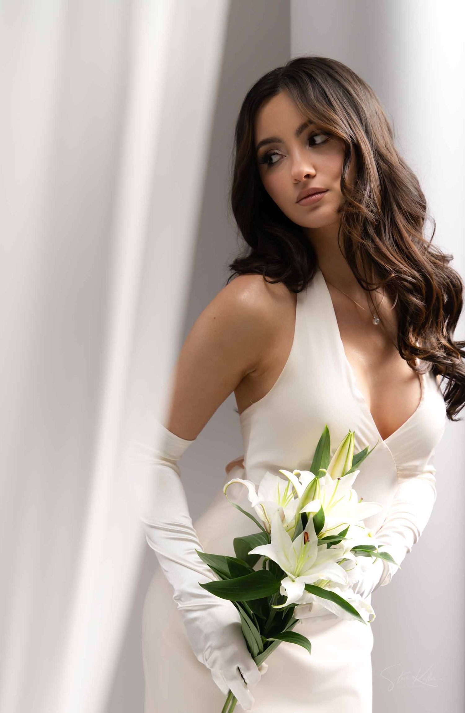 Woman in white dress holds white lily bouquet while posing against white curtain backdrop in elegant portrait series.