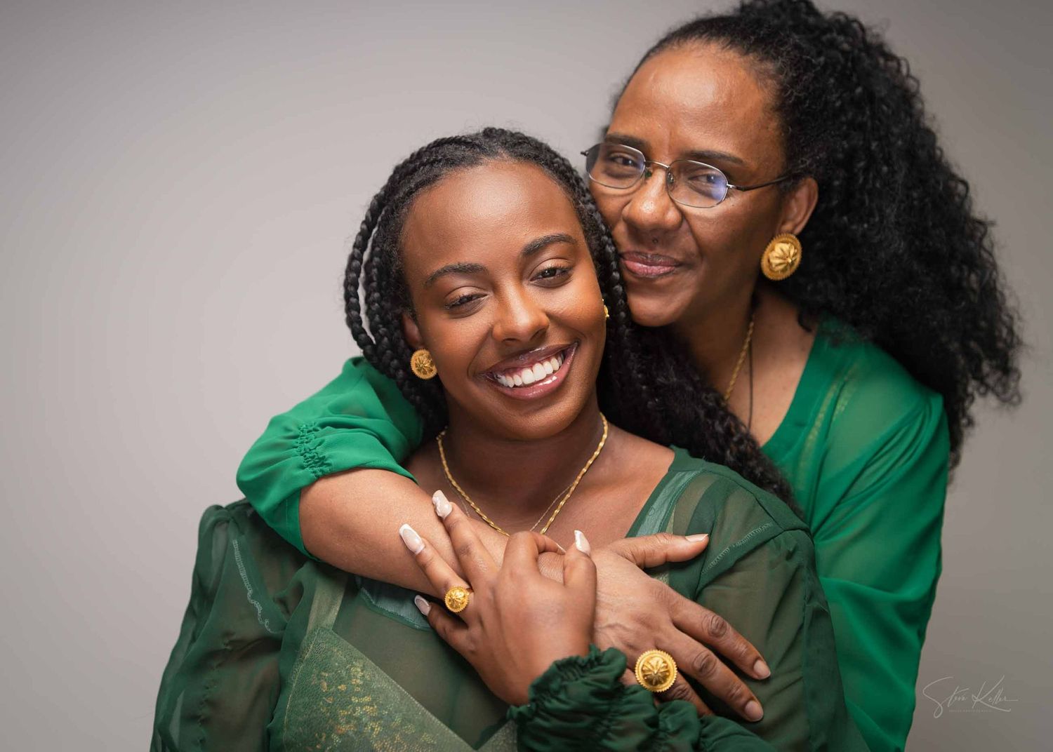 Two people wearing green tops embrace warmly while smiling against a gray background.