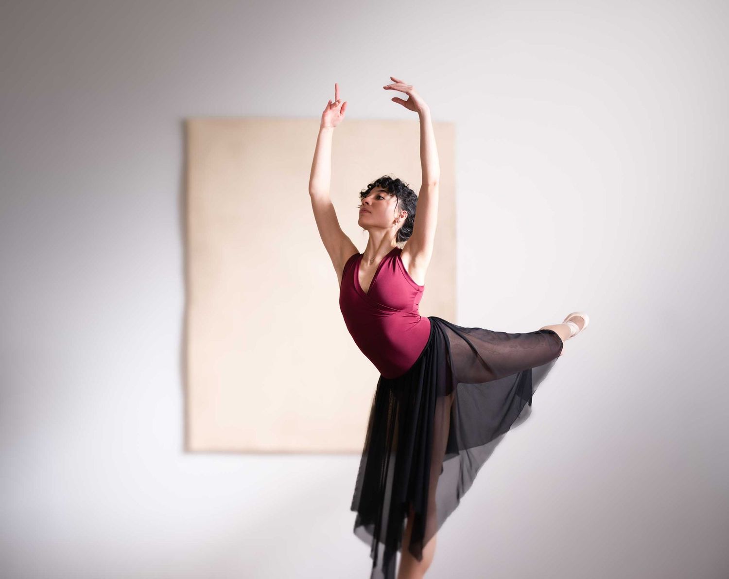 Graceful ballet dancer in red leotard and black skirt performs arabesque with arms raised overhead.