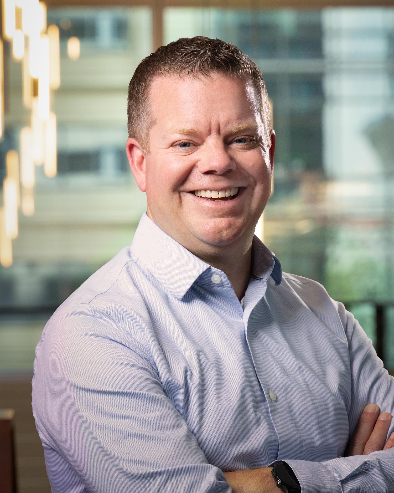 Professional business portrait of someone in a light blue dress shirt against a blurred office background.