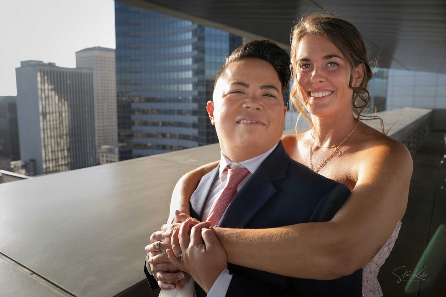 Newlywed couple poses together on sunny rooftop with city buildings in background.