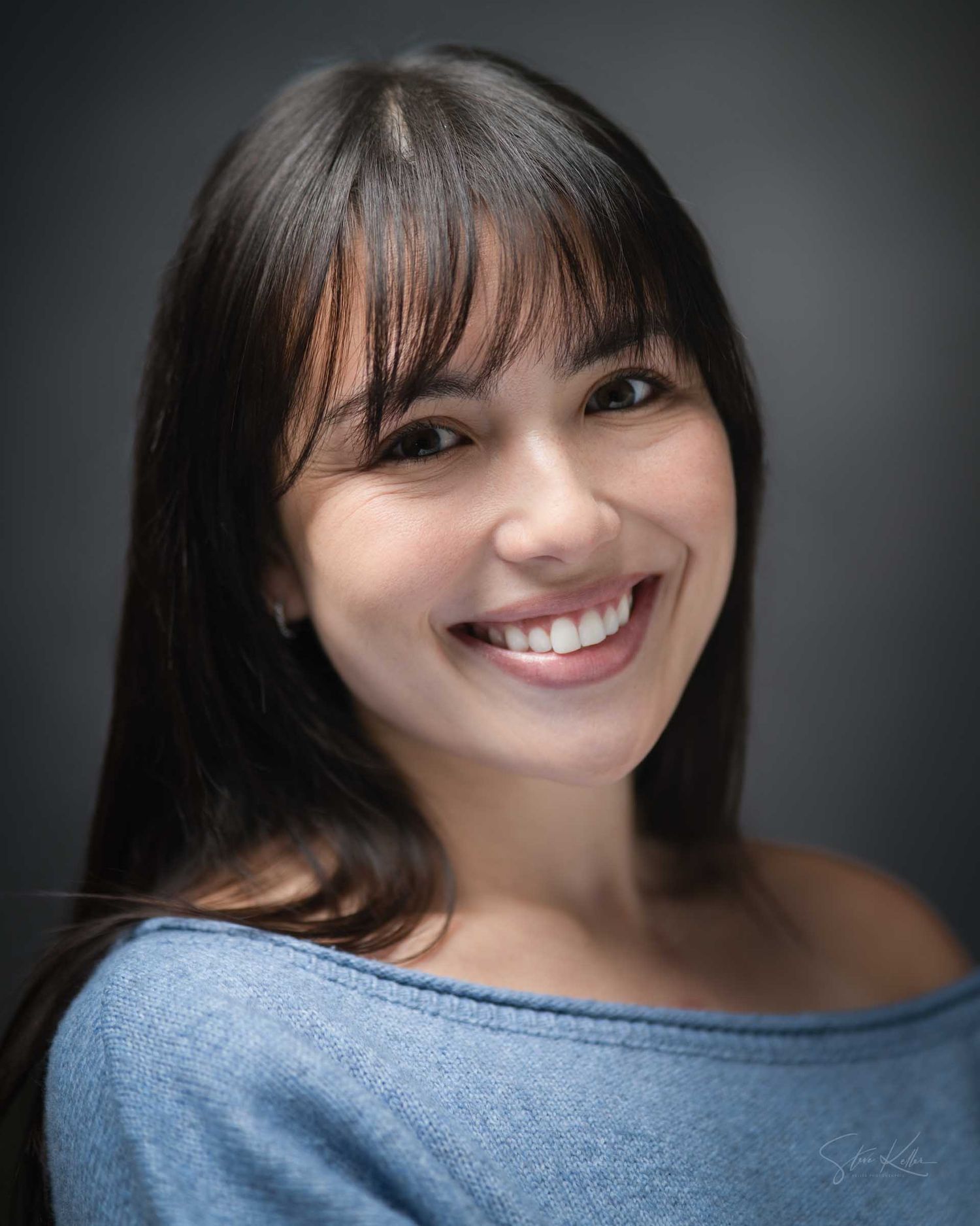 Smiling young woman with dark hair and bangs wearing a light blue off-shoulder top against a gray background.