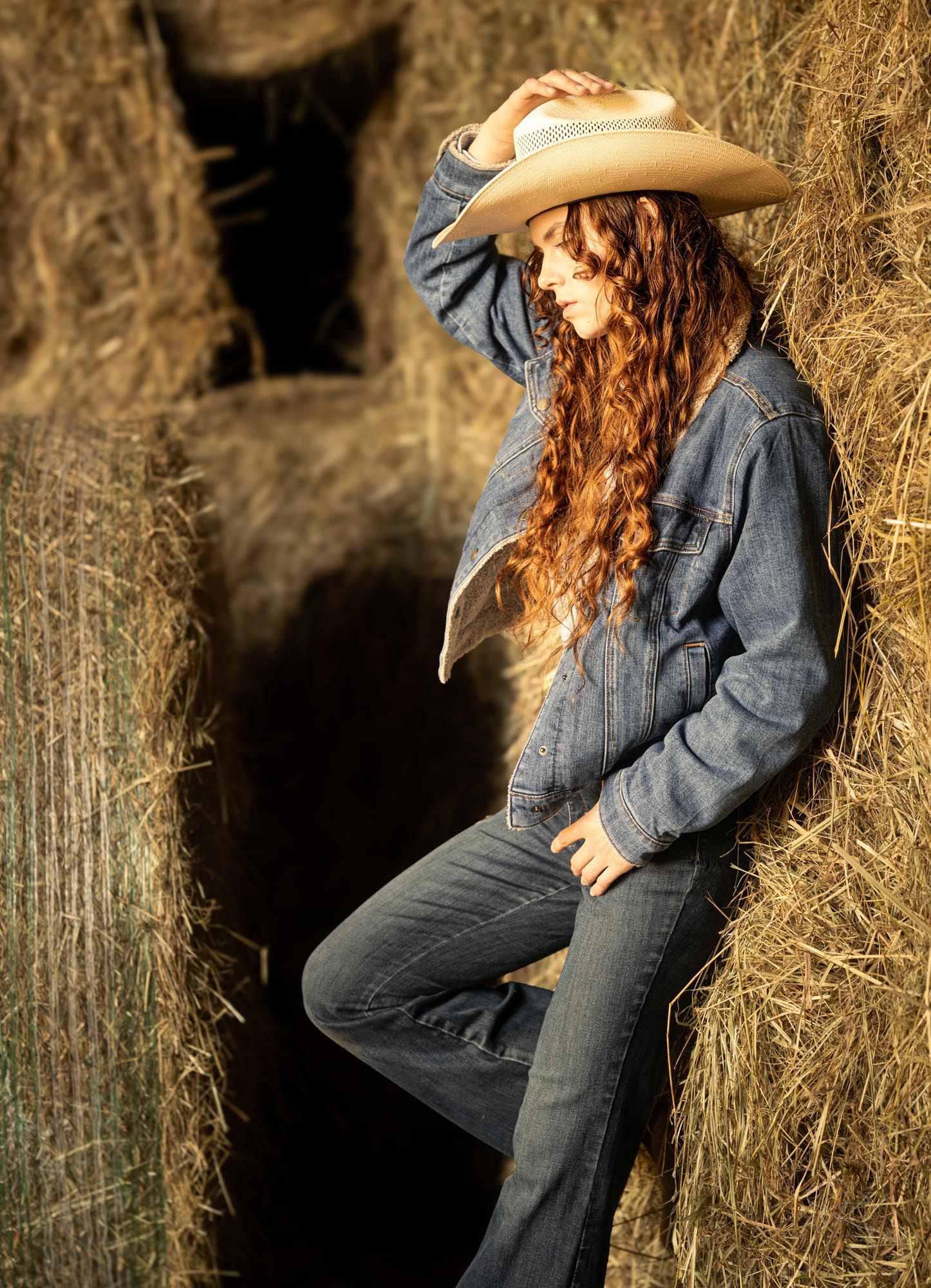 Young woman with long red hair wearing a denim jacket and cowboy hat posing against hay bales.