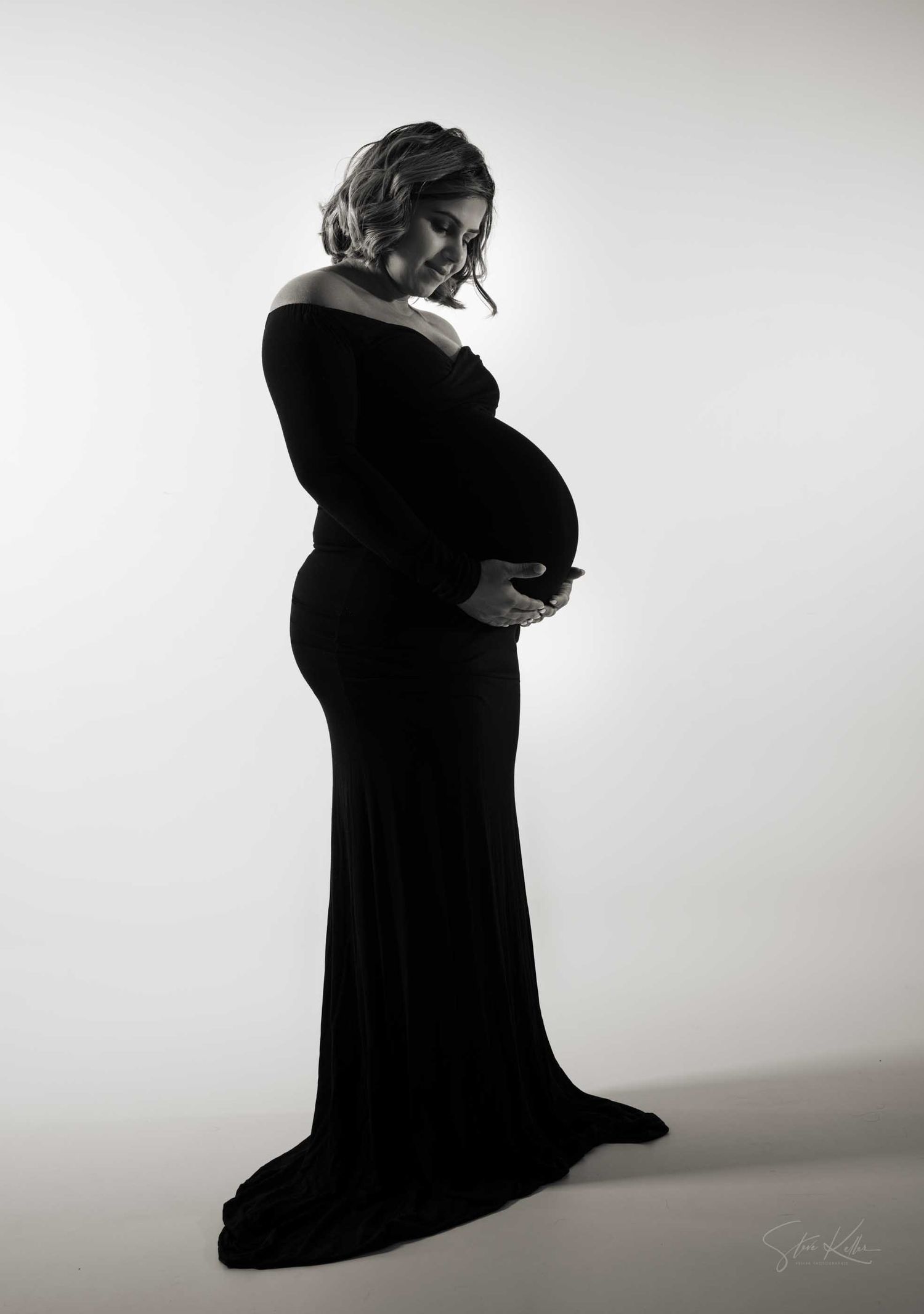 Black and white maternity photo of a pregnant woman in an elegant off-shoulder black gown, cradling her baby bump.