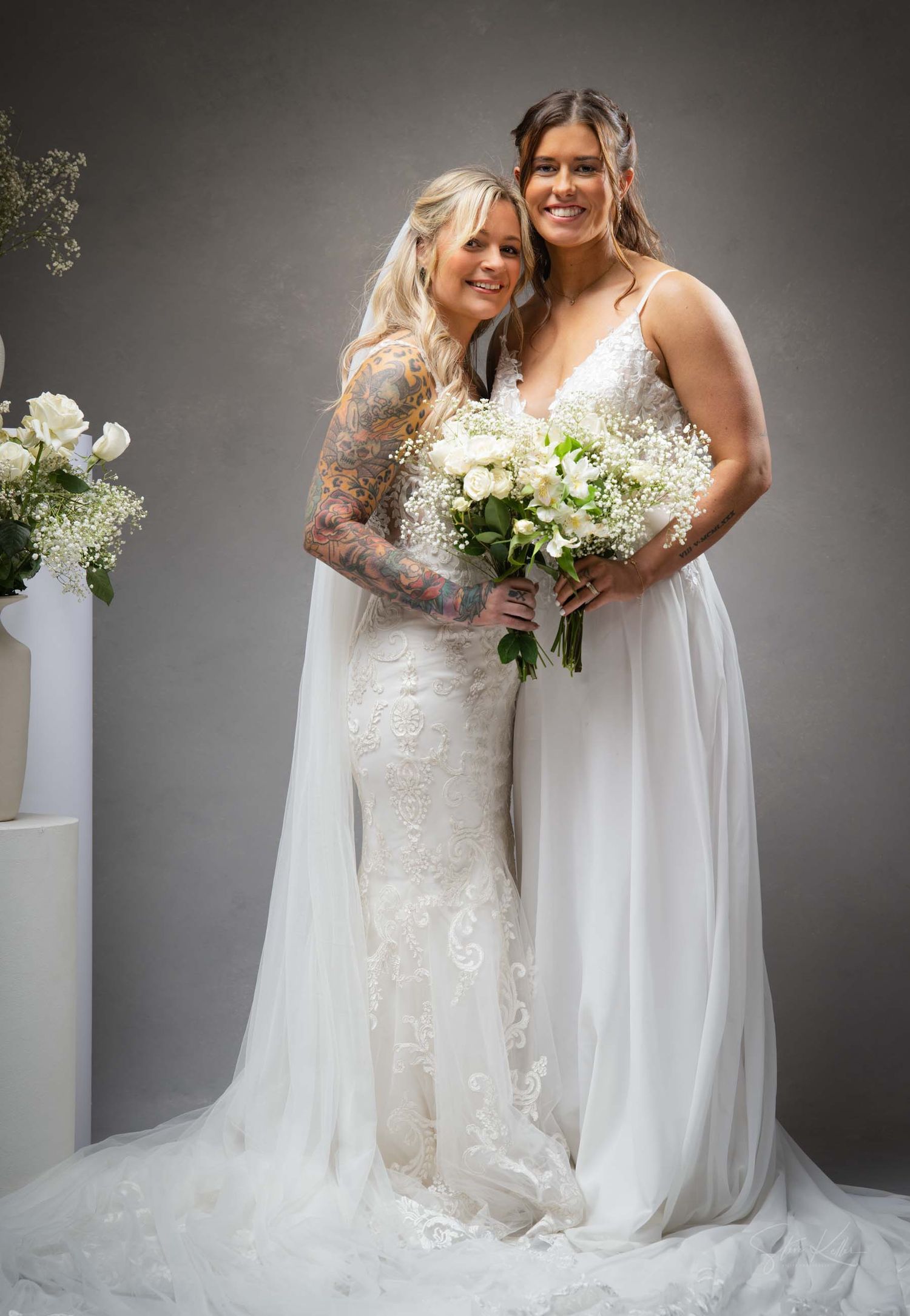 Two brides in flowing white gowns pose together against a grey backdrop holding matching bouquets.