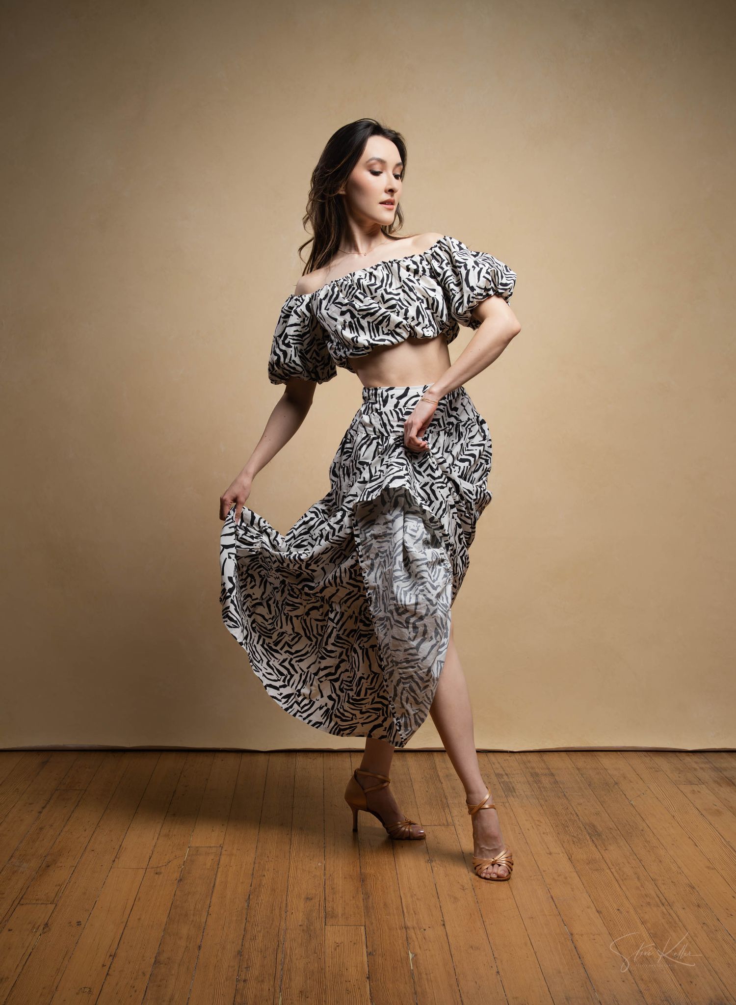 A dancer in a black and white patterned two-piece outfit twirls gracefully on a wooden floor against a neutral backdrop.