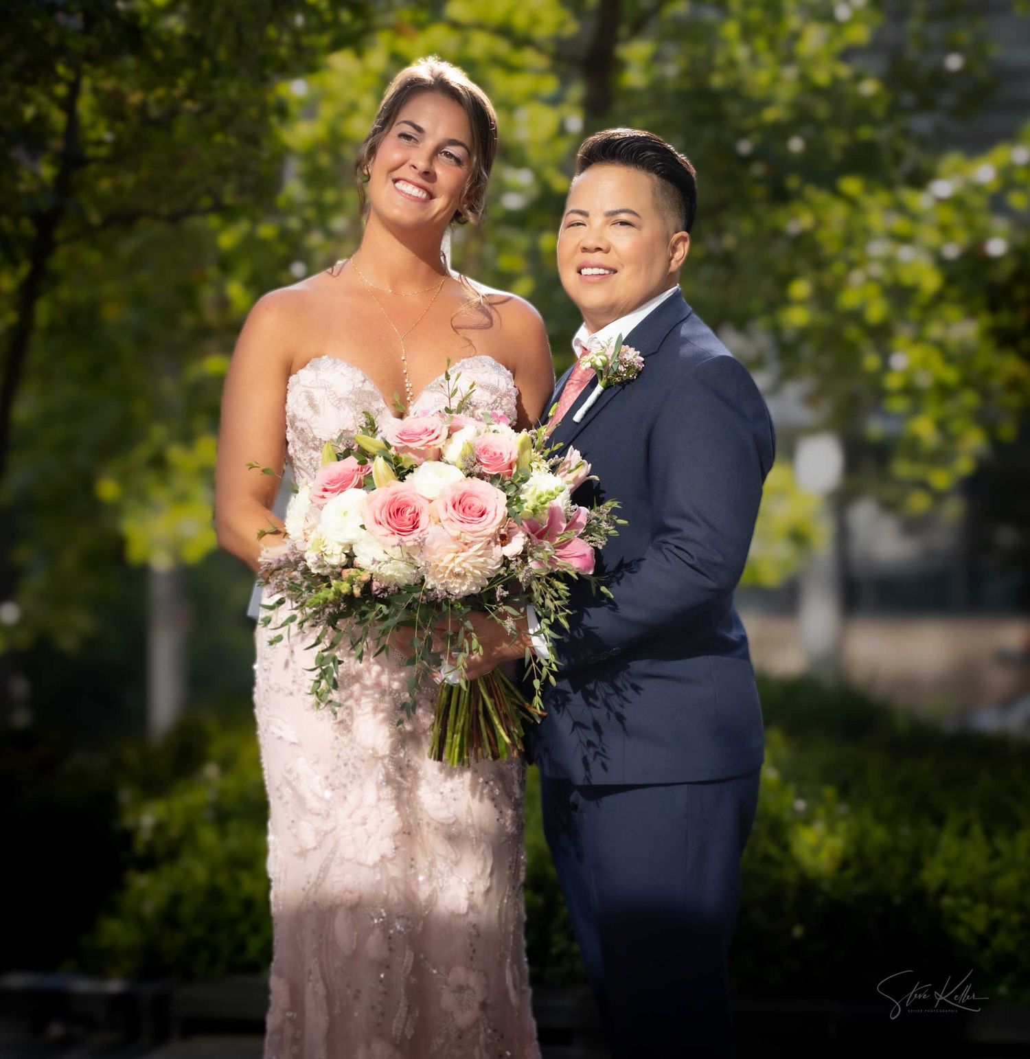 Two brides pose outdoors, one in a lace gown holding pink bouquet, other in navy suit, celebrating their wedding day.