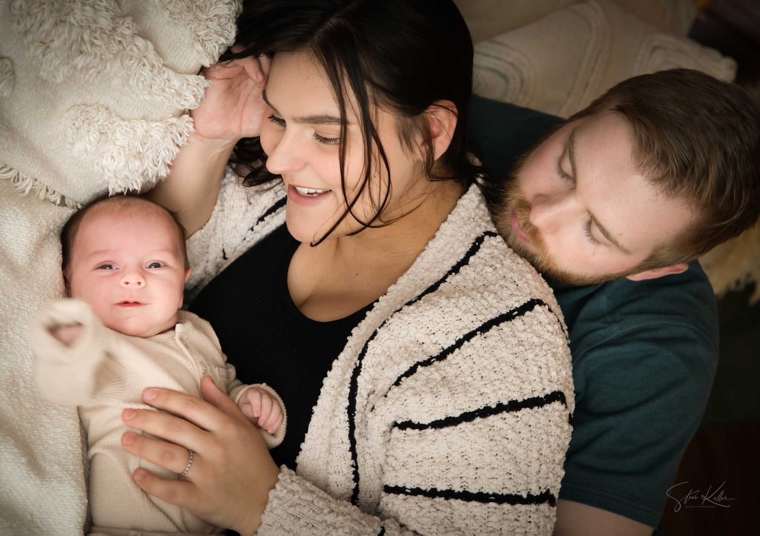 New parents lovingly gaze at their newborn baby while lying together on a cozy bed in warm light.