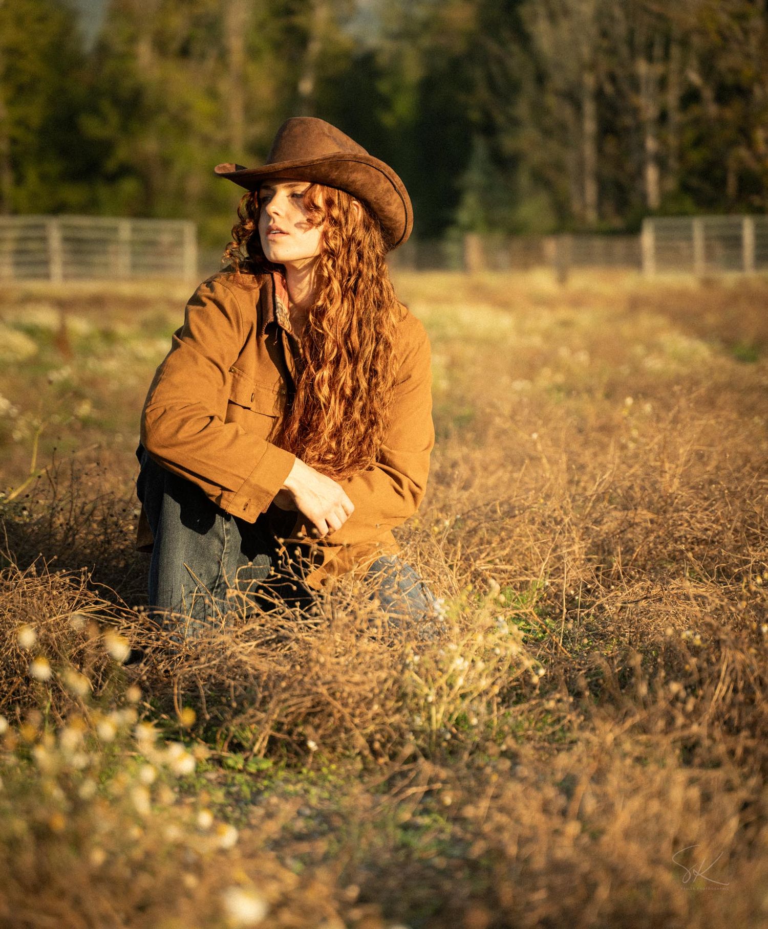 A young woman with long curly red hair wearing a brown jacket and cowboy hat sits in a dry field.