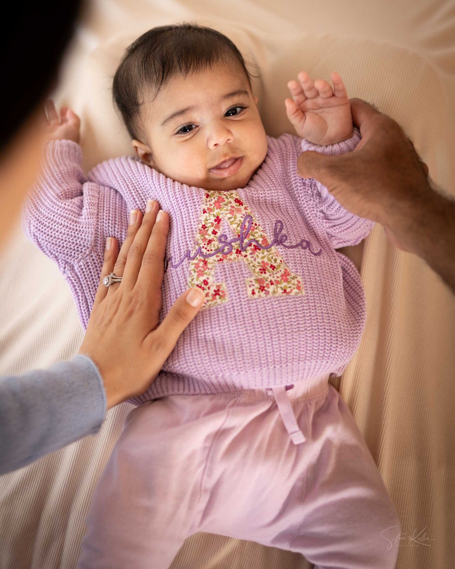 Adorable baby in purple knit sweater with floral embroidery lies on cream blanket held by parents.