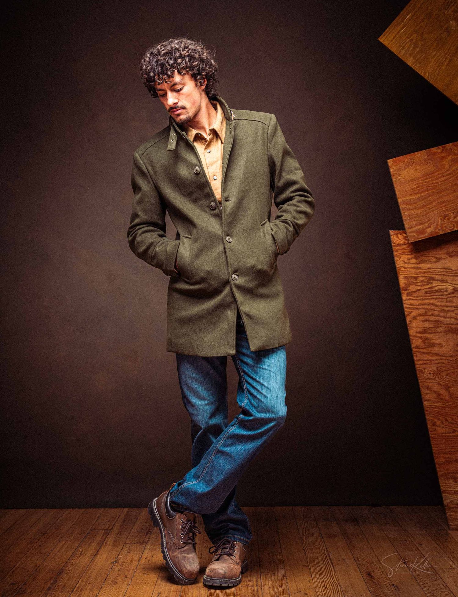 A person in a green jacket and blue jeans stands on a wooden floor in an artistic dance-like pose against a dark backdrop.