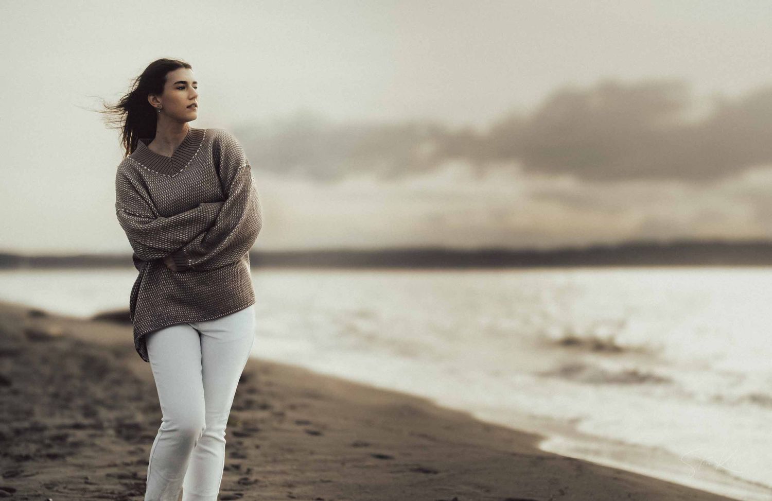 Person in cozy sweater walking along misty beach shoreline at sunset in serene coastal setting.