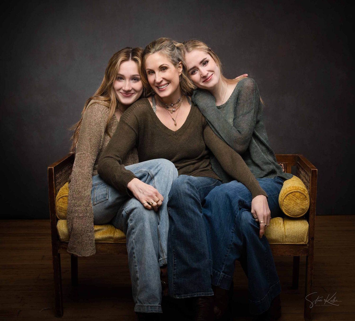 Three people sit close together on a brown couch in a studio setting with dark background.