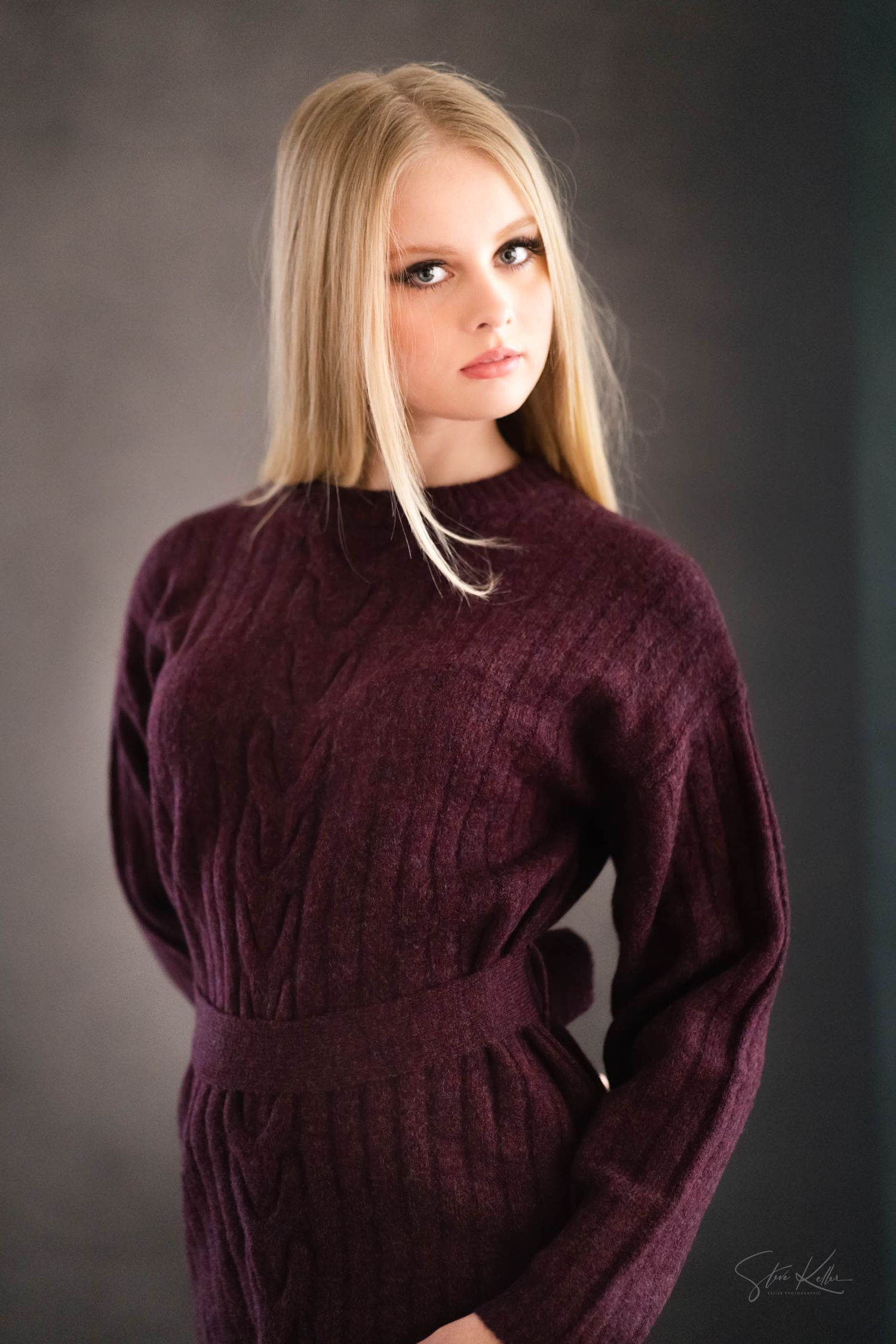 Young blonde woman in a burgundy cable-knit sweater dress poses gracefully against a gray studio background.
