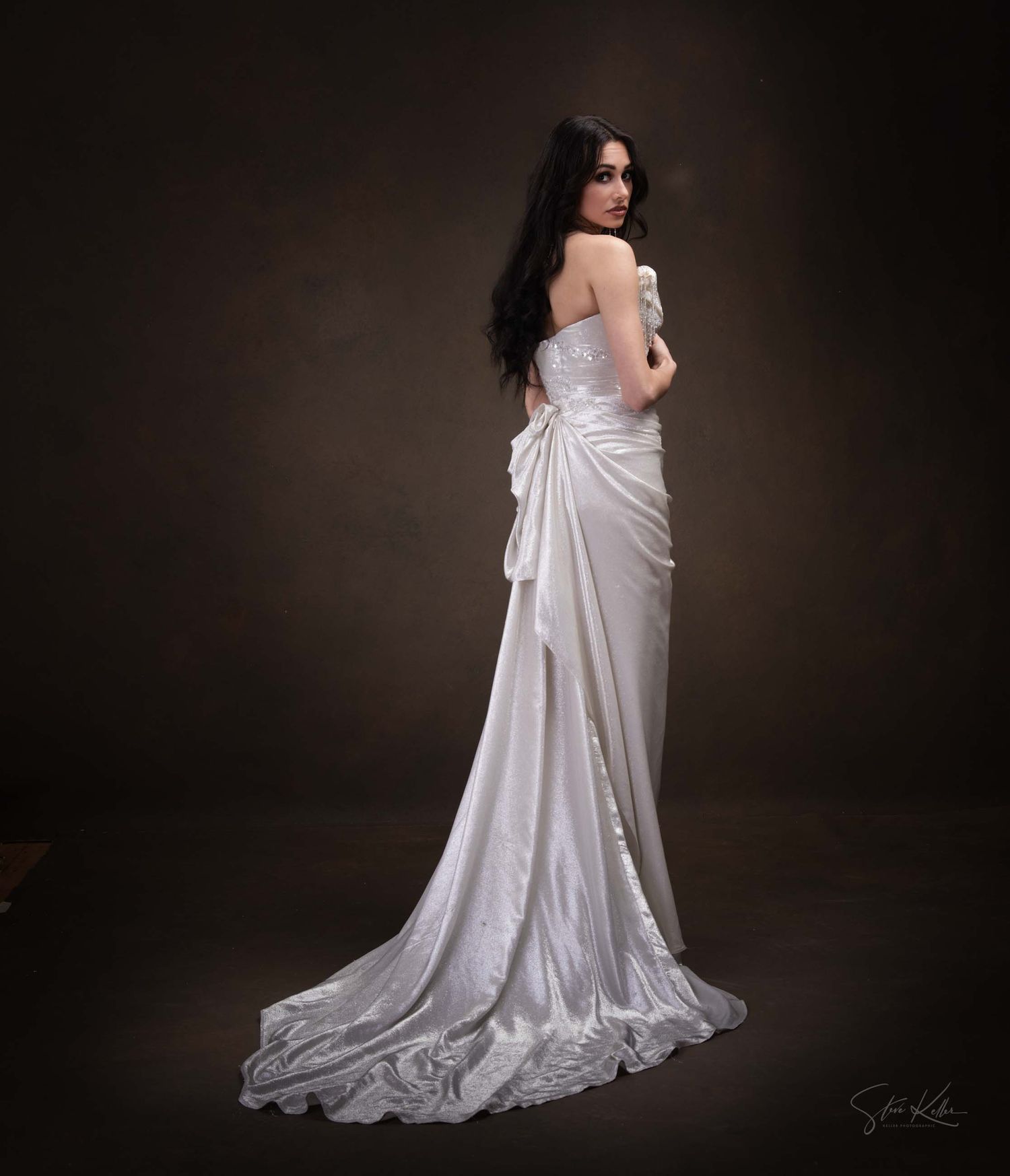 A bride in a flowing white satin wedding gown with train poses elegantly against a dark studio backdrop.