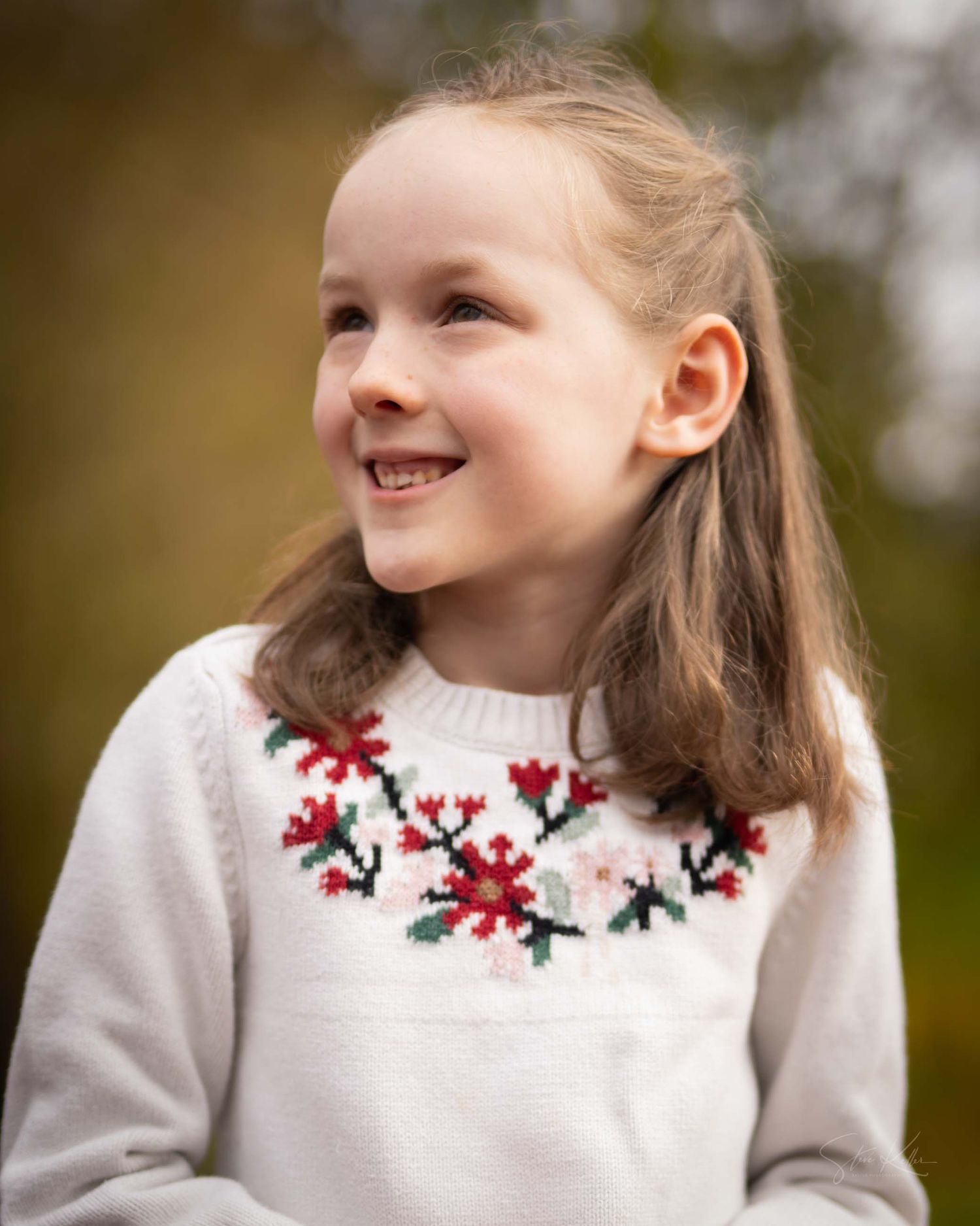 A child wearing a white sweater with red floral embroidery smiles while looking up and away from the camera.