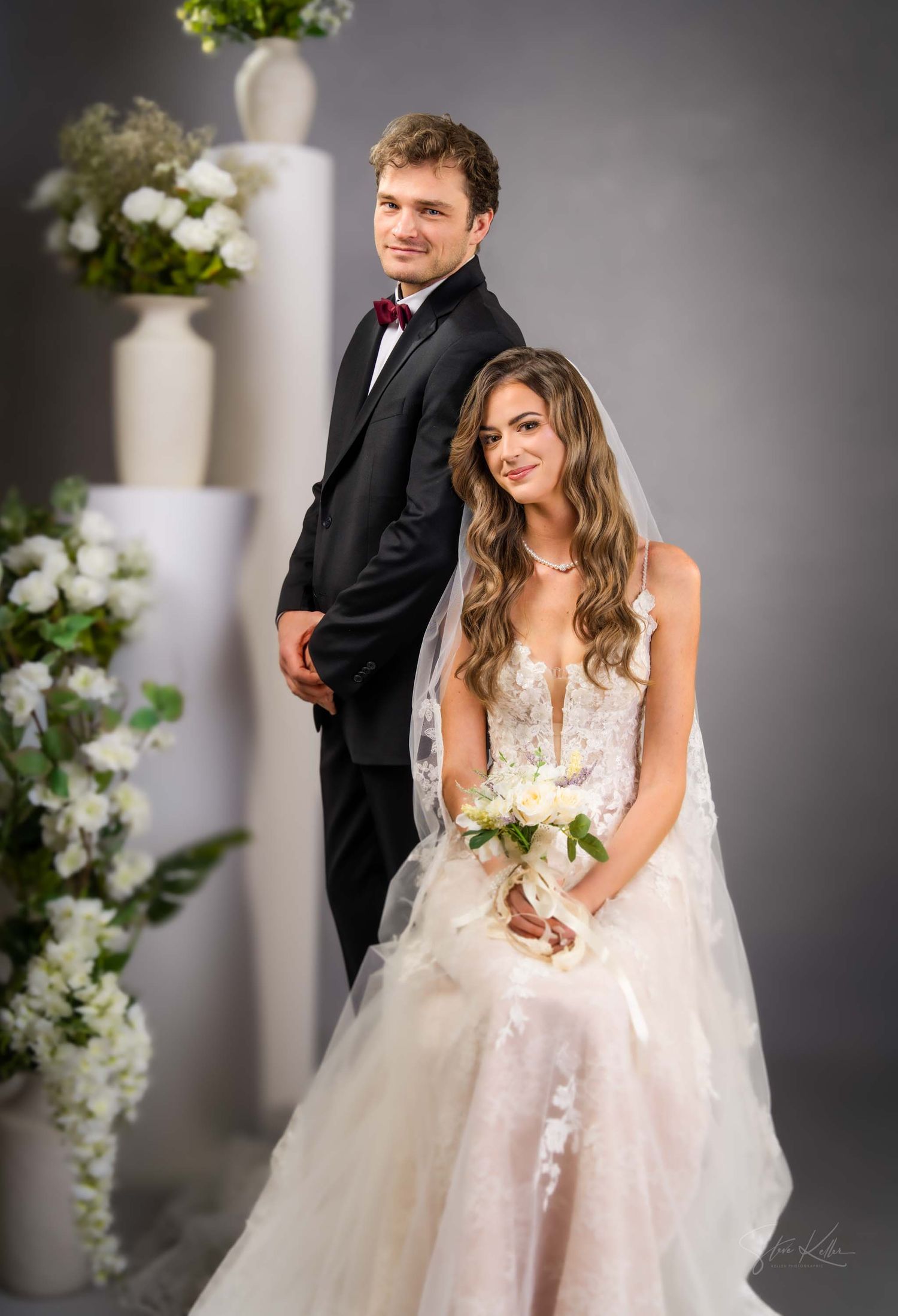 A bride in a lace wedding gown and groom in black tuxedo pose together among white floral arrangements.