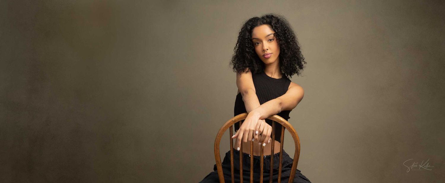 Young woman with curly hair poses elegantly on a wooden chair against a gray studio background.