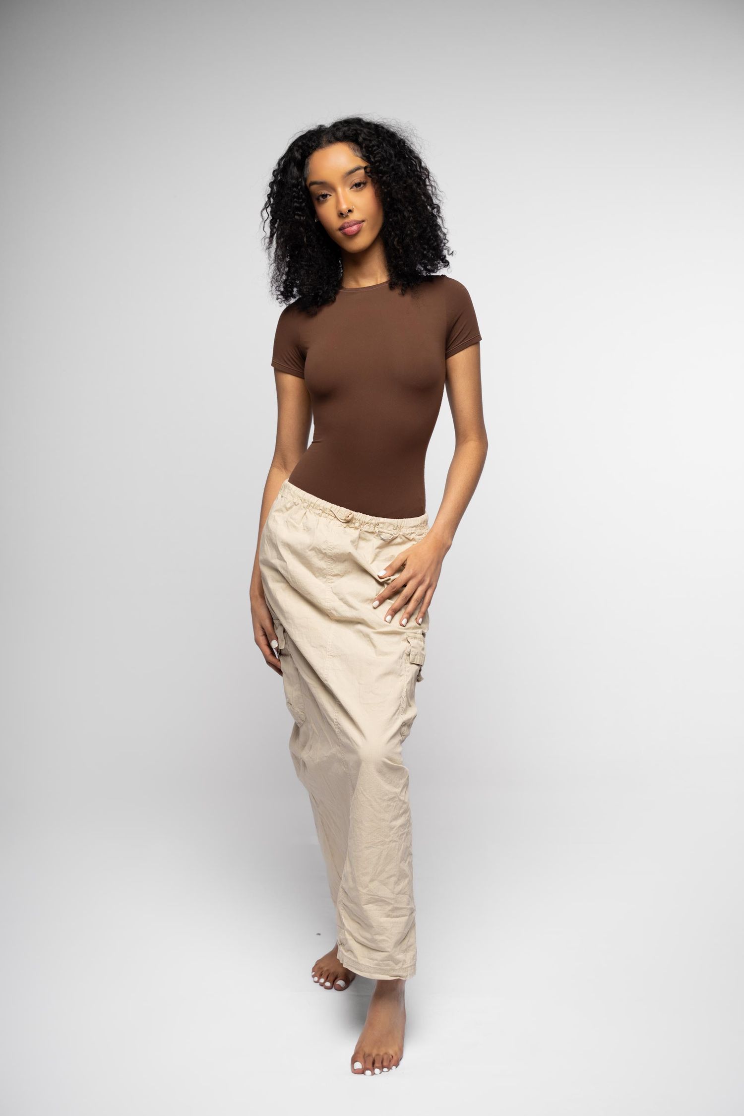 Fashion model in brown fitted t-shirt and beige cargo pants poses barefoot against light gray studio background.