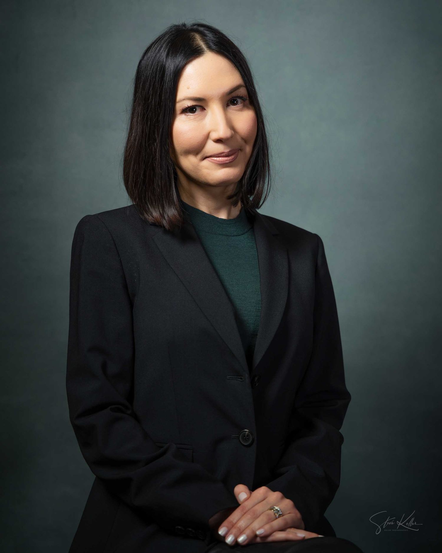 Professional headshot of a woman in a black blazer and teal top, with dark hair, posing against a gray background.
