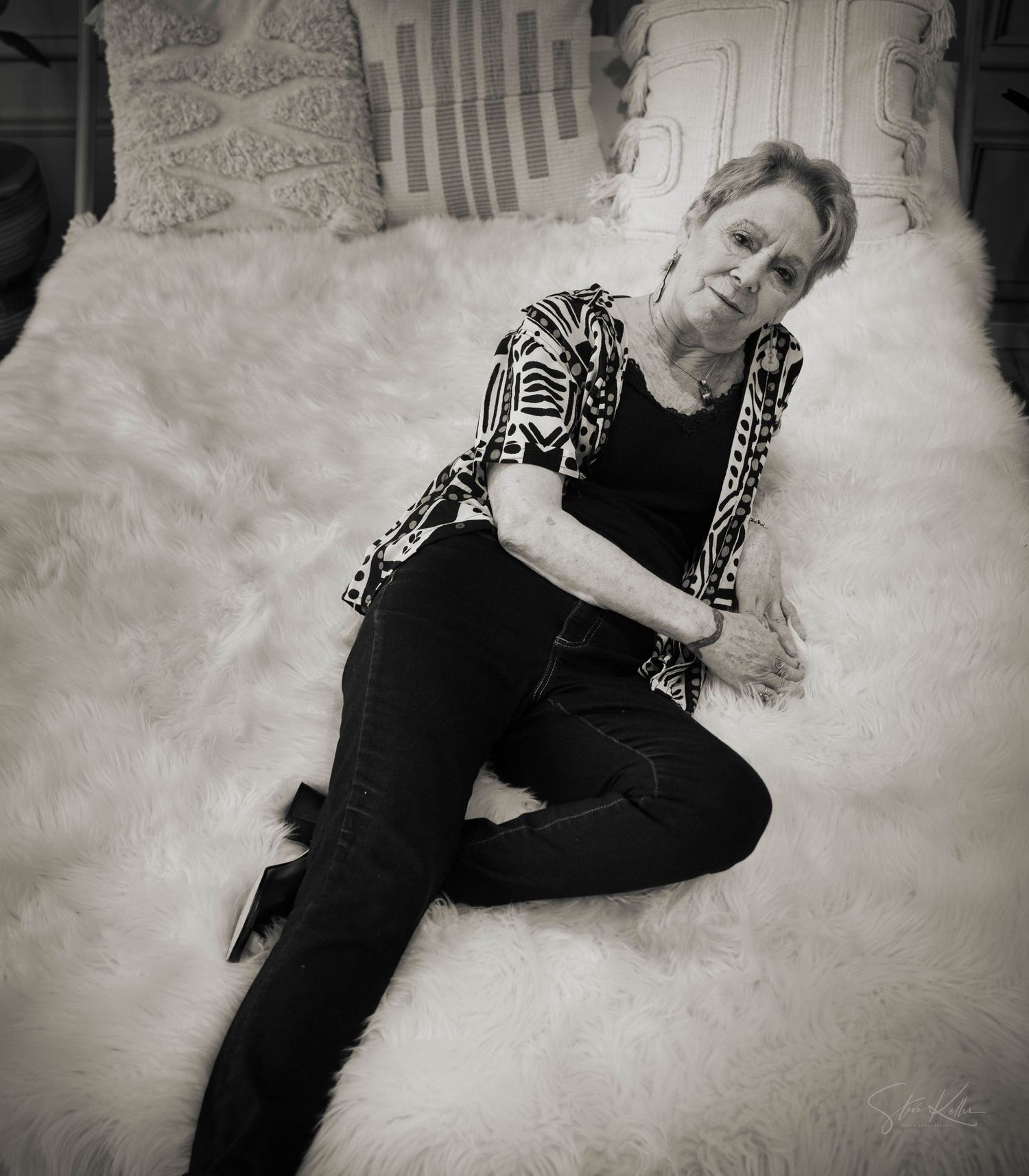 Black and white portrait of someone lounging on white fur wearing a patterned top and dark pants.