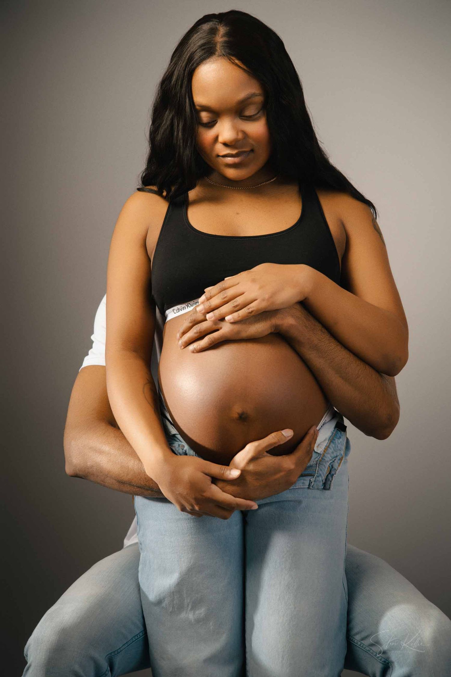 A series of maternity photos showing a pregnant person in denim jeans cradling their belly against a neutral backdrop.
