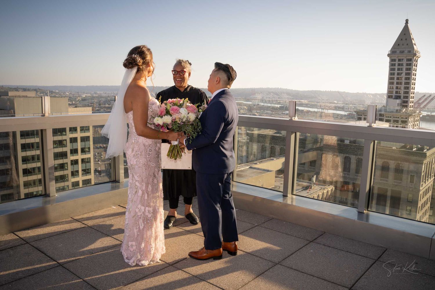 Bride and groom stand with officiant during rooftop wedding ceremony at golden hour.