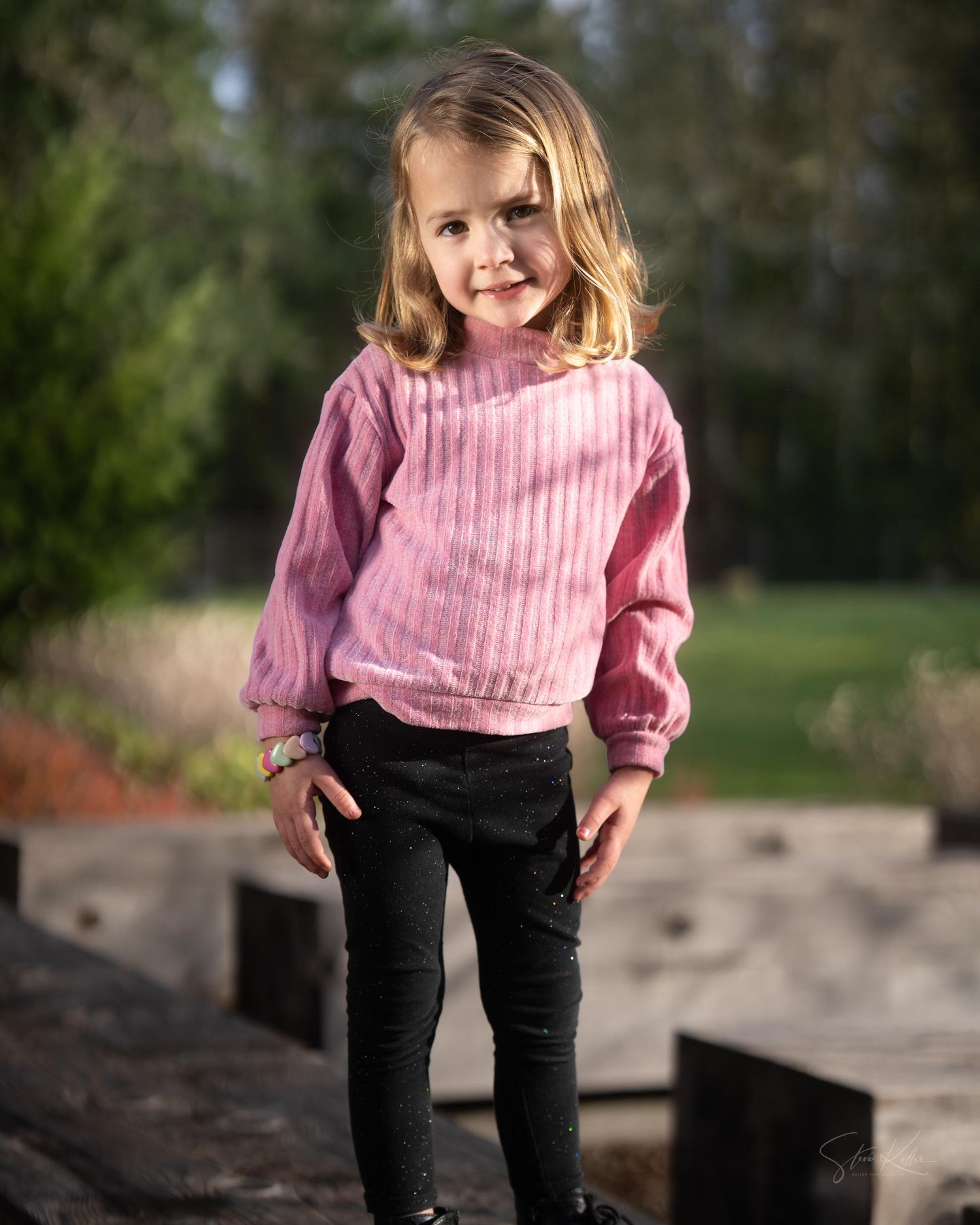 Child wearing a pink striped sweater and black pants plays outdoors on a sunny day.