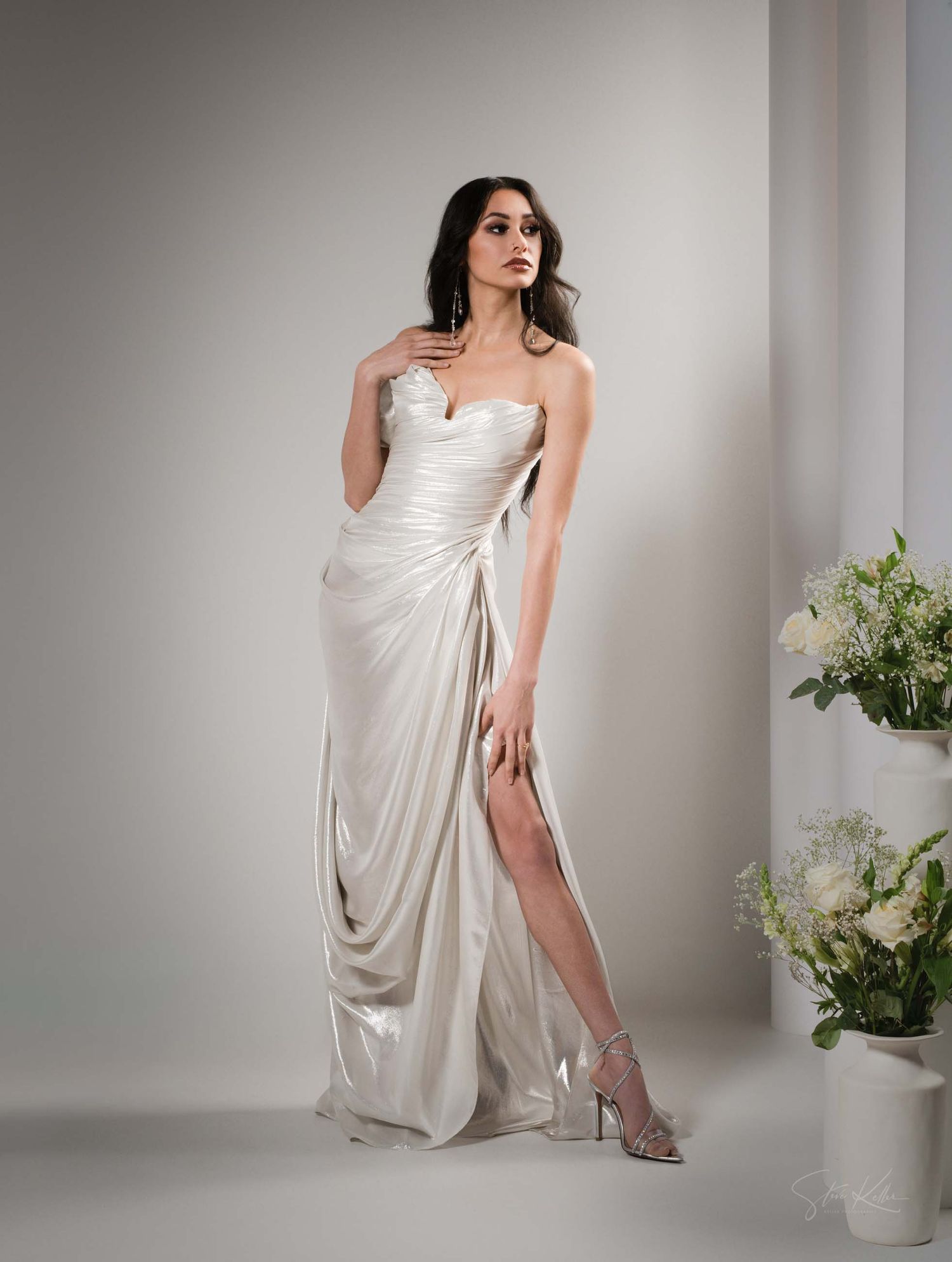 Elegant strapless ivory satin wedding gown with draped fabric, thigh-high slit and floral decor in a studio setting.