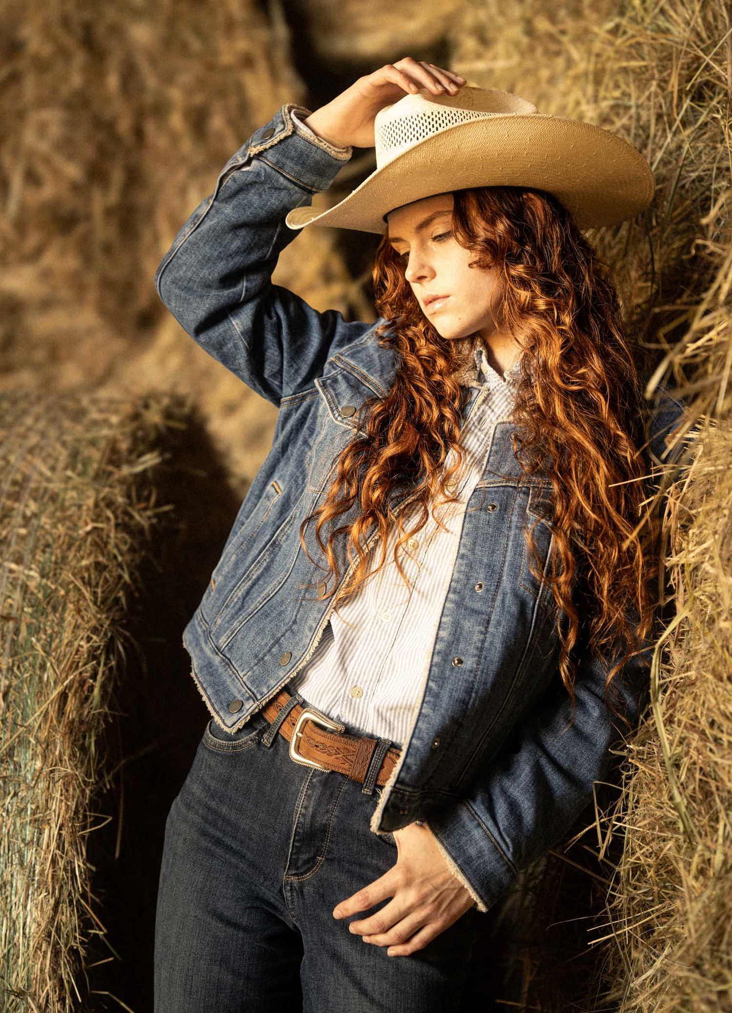 Woman with long red curly hair wearing a cowboy hat, denim jacket, and jeans leaning against hay bales.