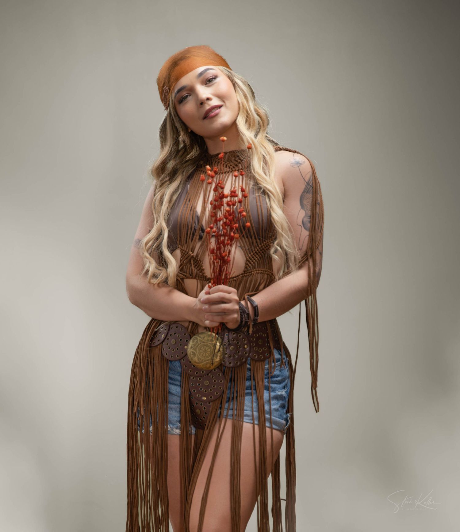 A bohemian-style fashion model in fringe leather vest and denim shorts poses against neutral background.