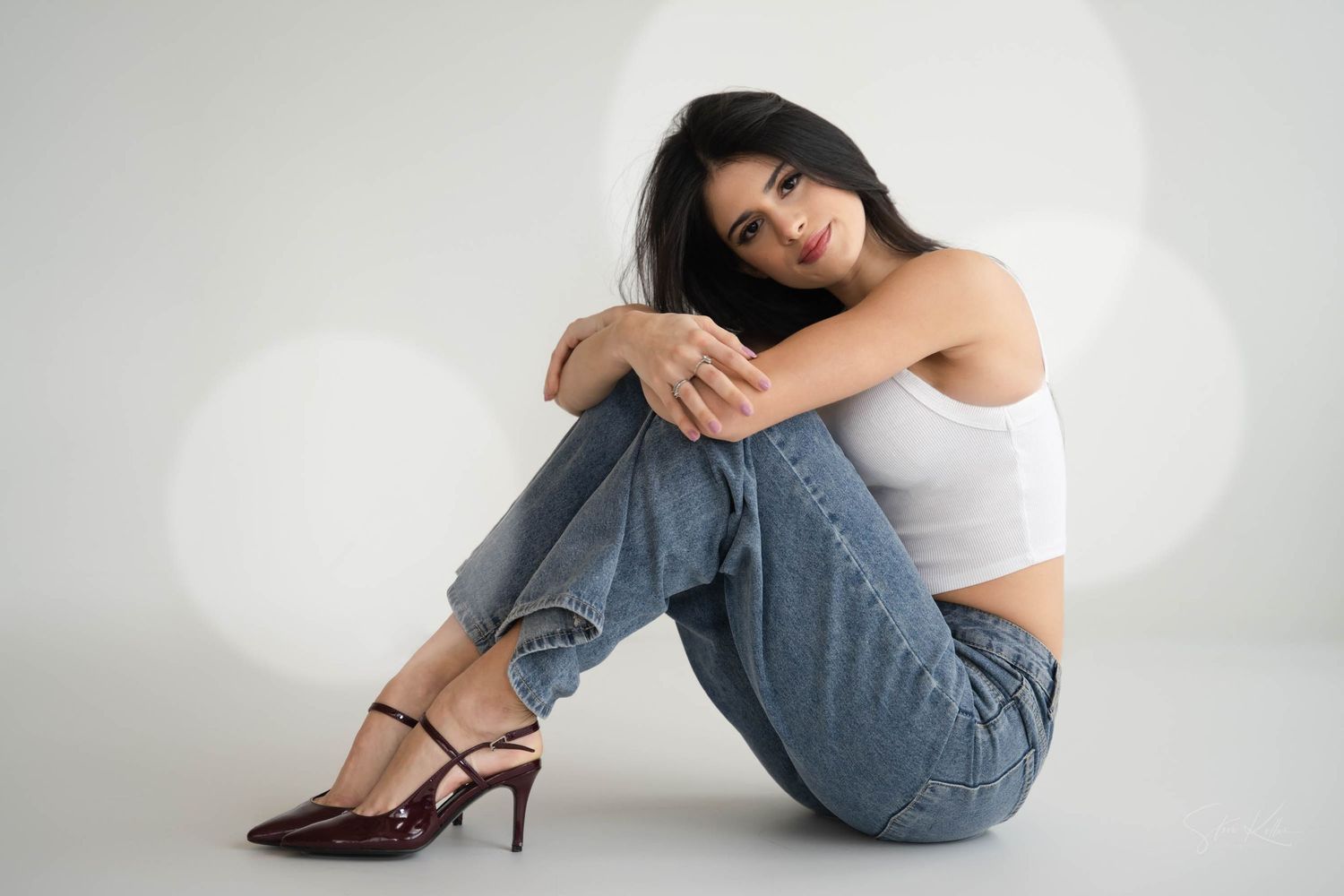 fashion model in white crop top and light blue jeans sits casually on white floor wearing brown high heel sandals.