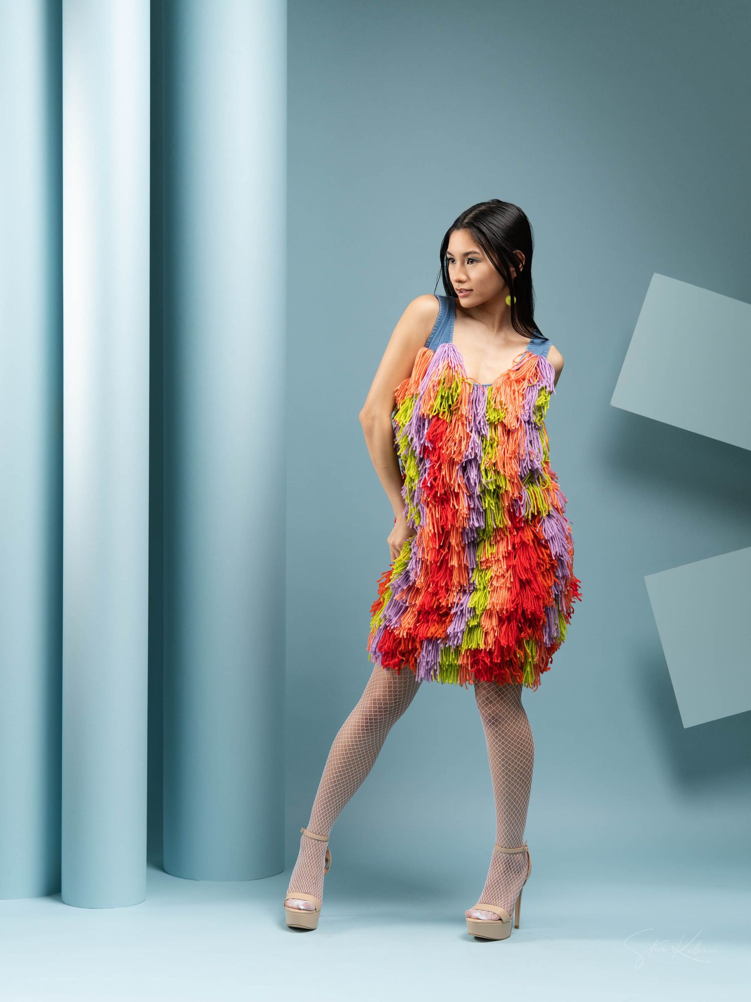 Model wearing vibrant multicolored fringe party dress with high heels poses against blue-gray studio background.