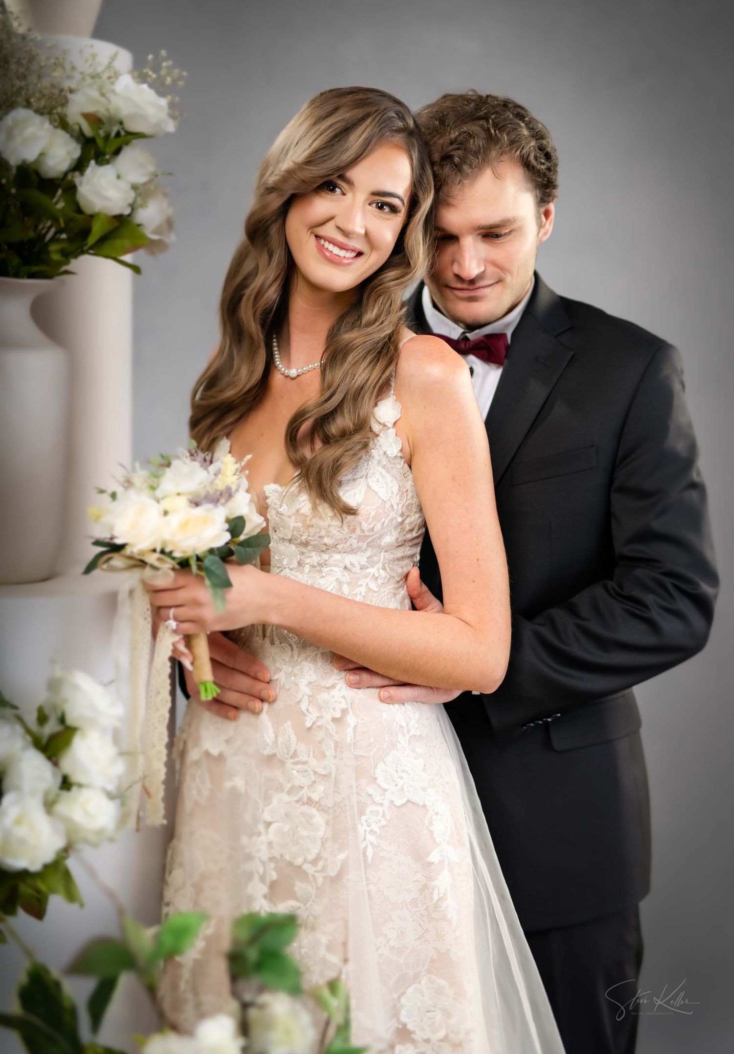 A couple in formal wedding attire pose together with white floral arrangements in an elegant studio setting.
