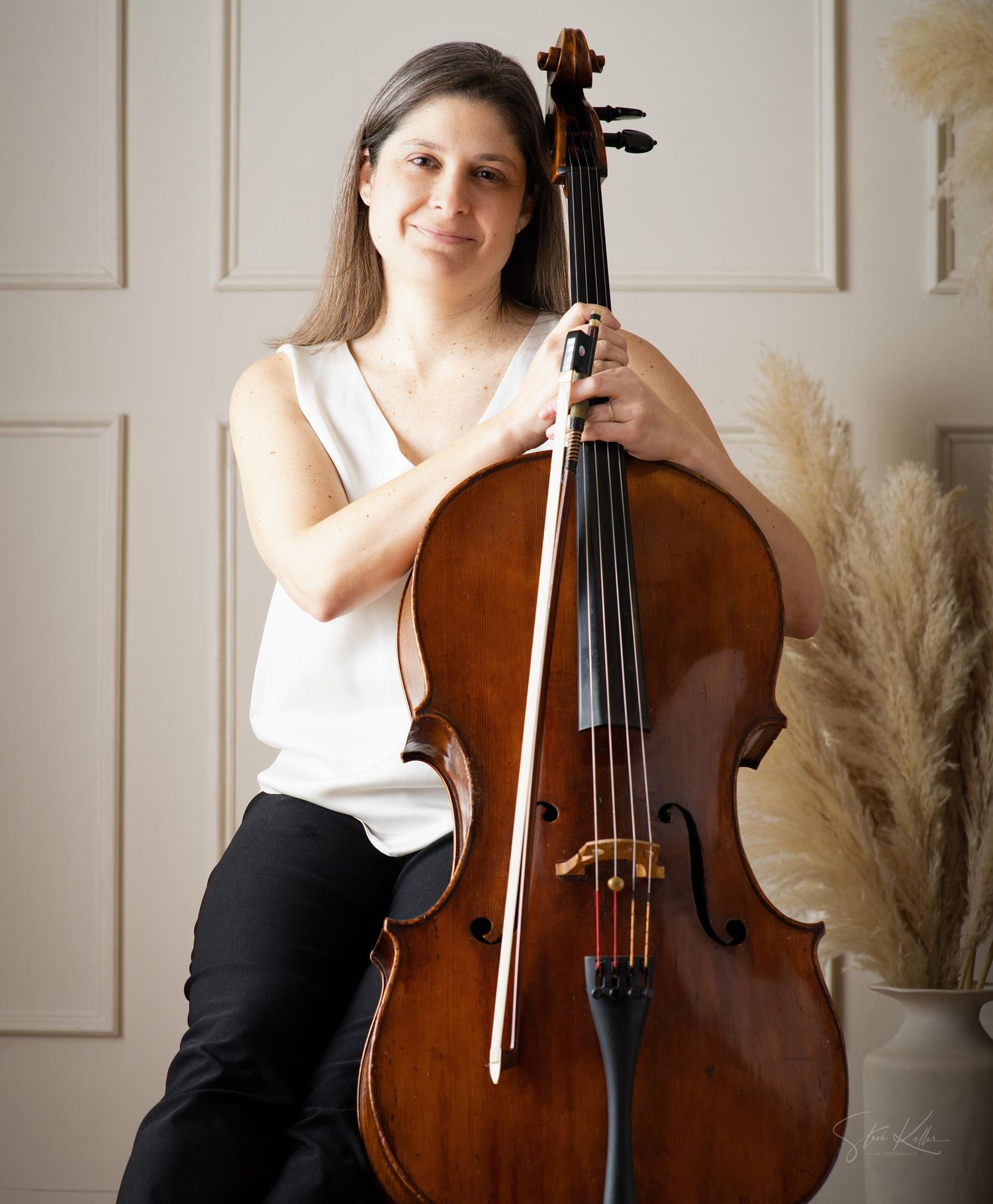 A musician poses with a wooden cello against a light wall with pampas grass decor in a bright studio setting.