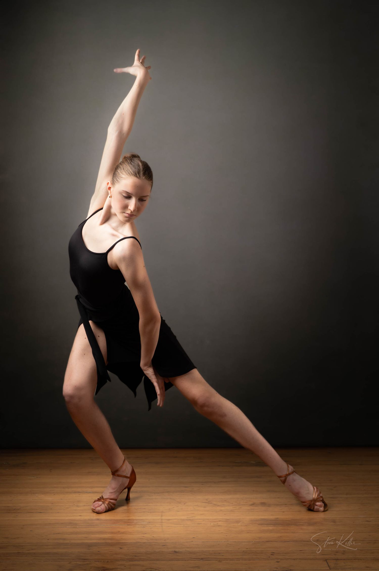 Dancer in black leotard strikes dramatic pose with outstretched arm on wooden dance floor against dark background.