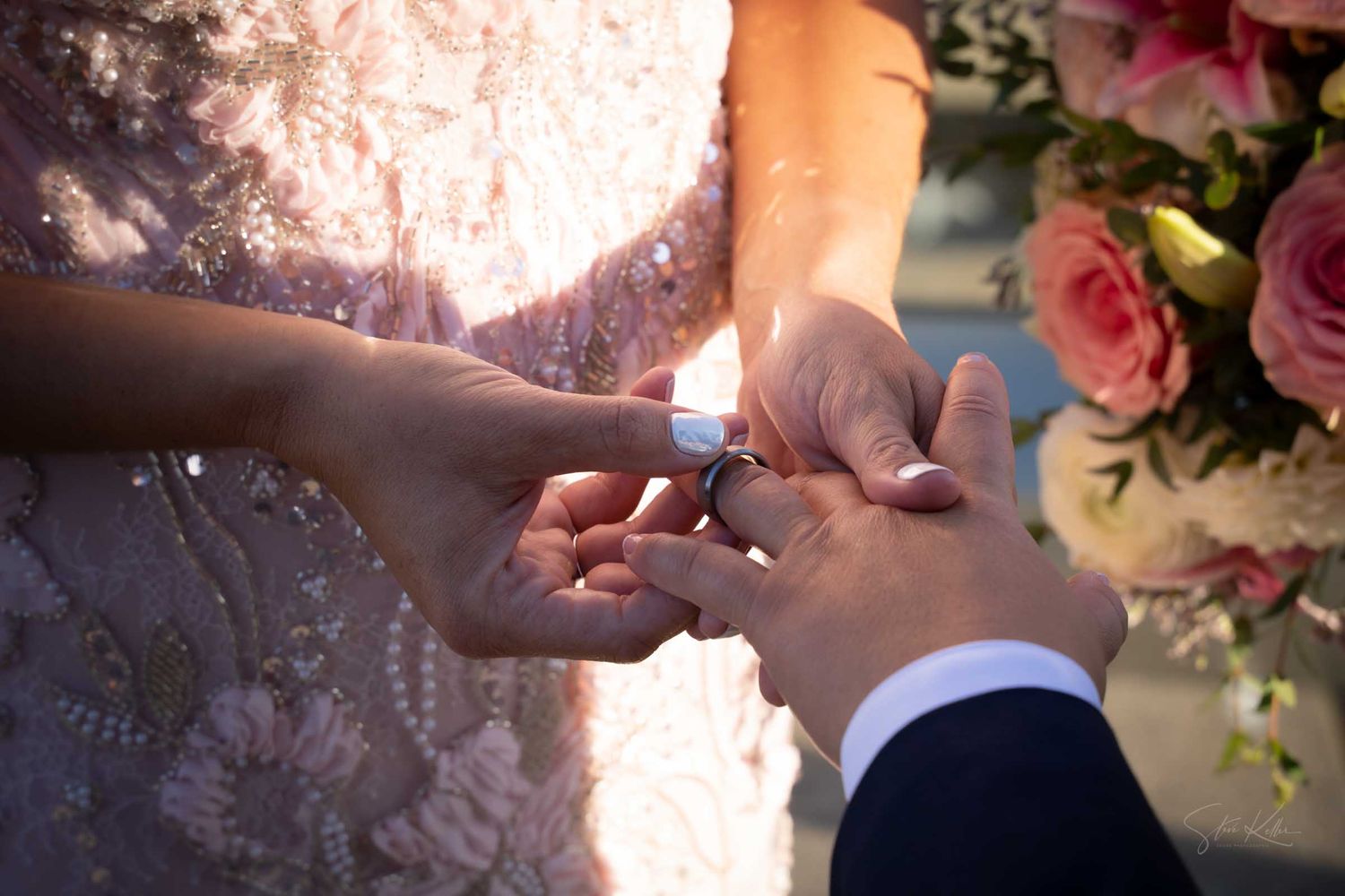 Close-up of a wedding ring exchange ceremony, with one bride in a pink lace gown placing a ring on her partners hand near pink roses.