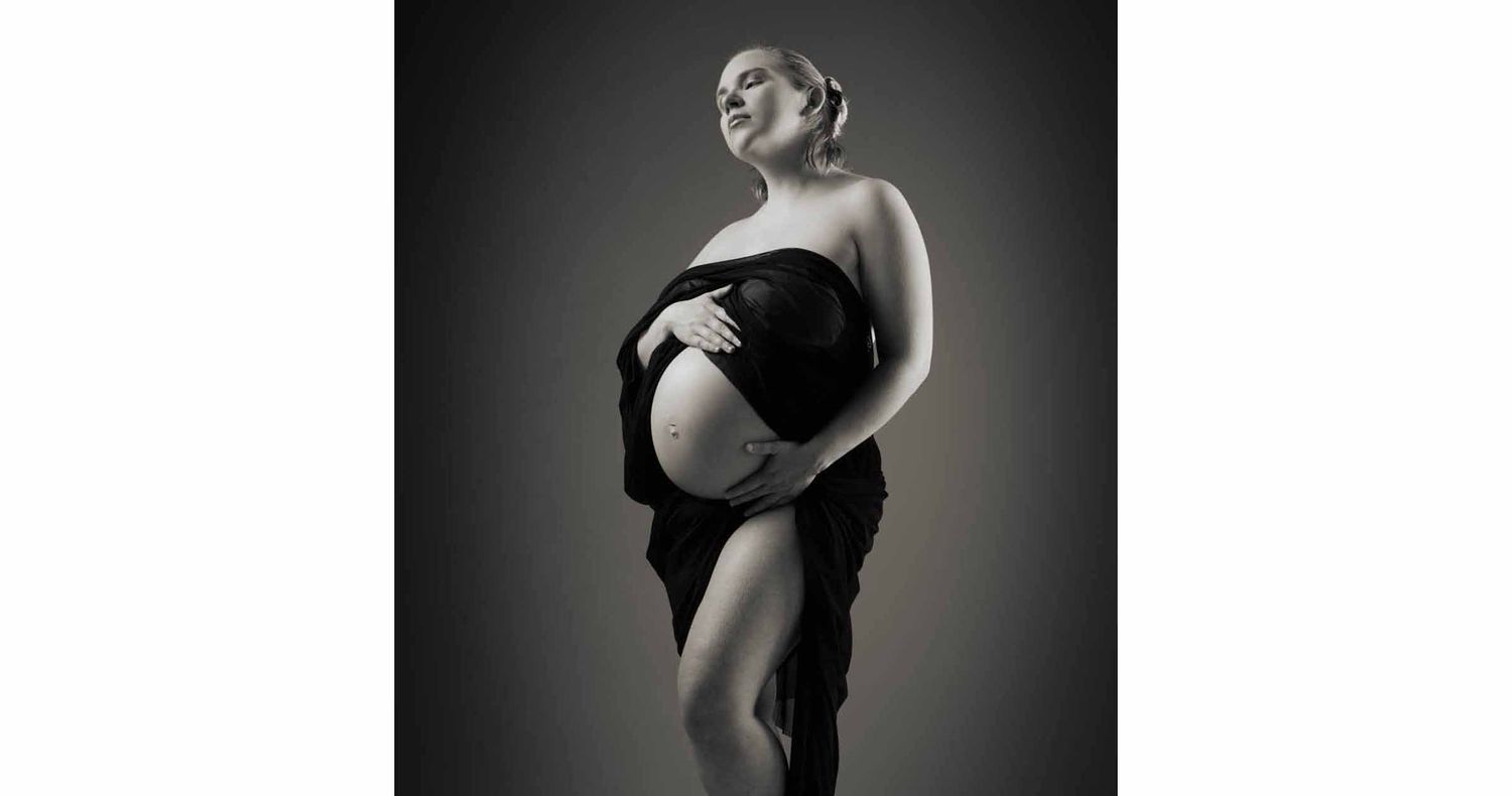Black and white maternity portrait of a pregnant woman draped in dark fabric, gently cradling her bare belly in a studio.