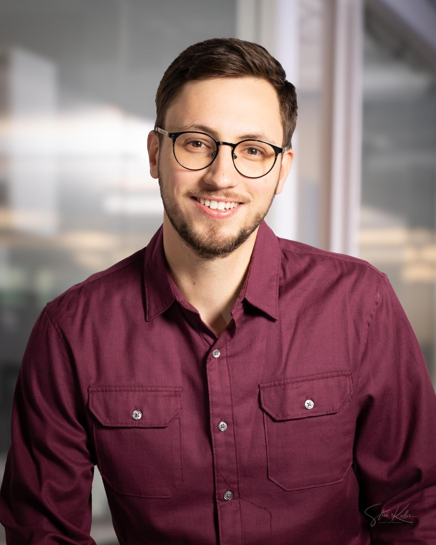 Professional headshot of person in burgundy button-up shirt with glasses against blurred office background.