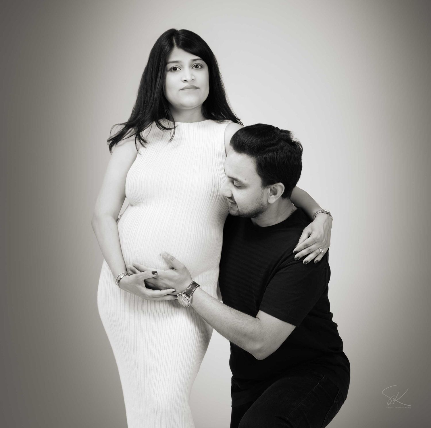 Black and white maternity photo of a pregnant woman in white dress with partner kissing her baby bump.