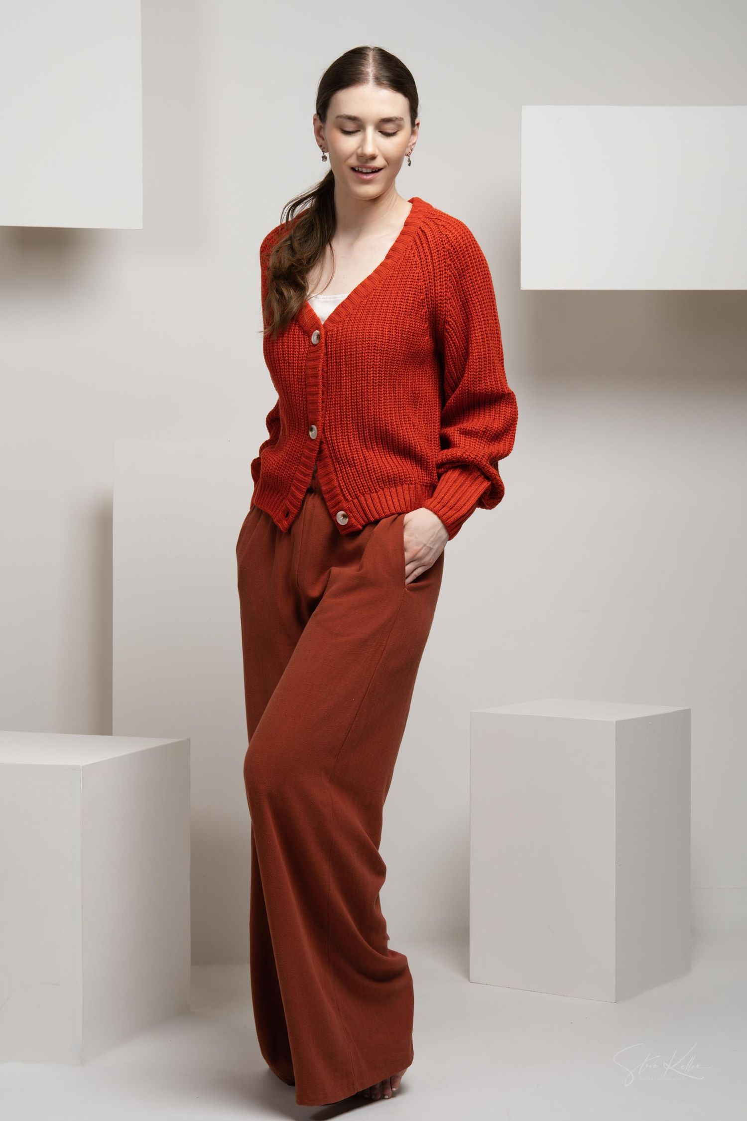 Model wearing a rust-colored knit cardigan and coordinating wide-leg pants in a minimal studio setting with white blocks.