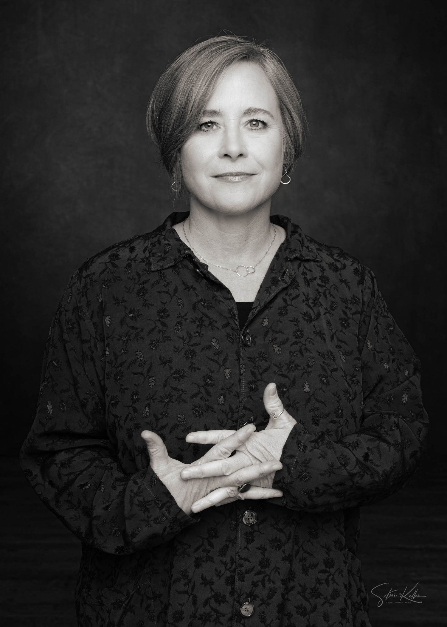 Professional studio portrait of woman in dark patterned blouse against dark background.