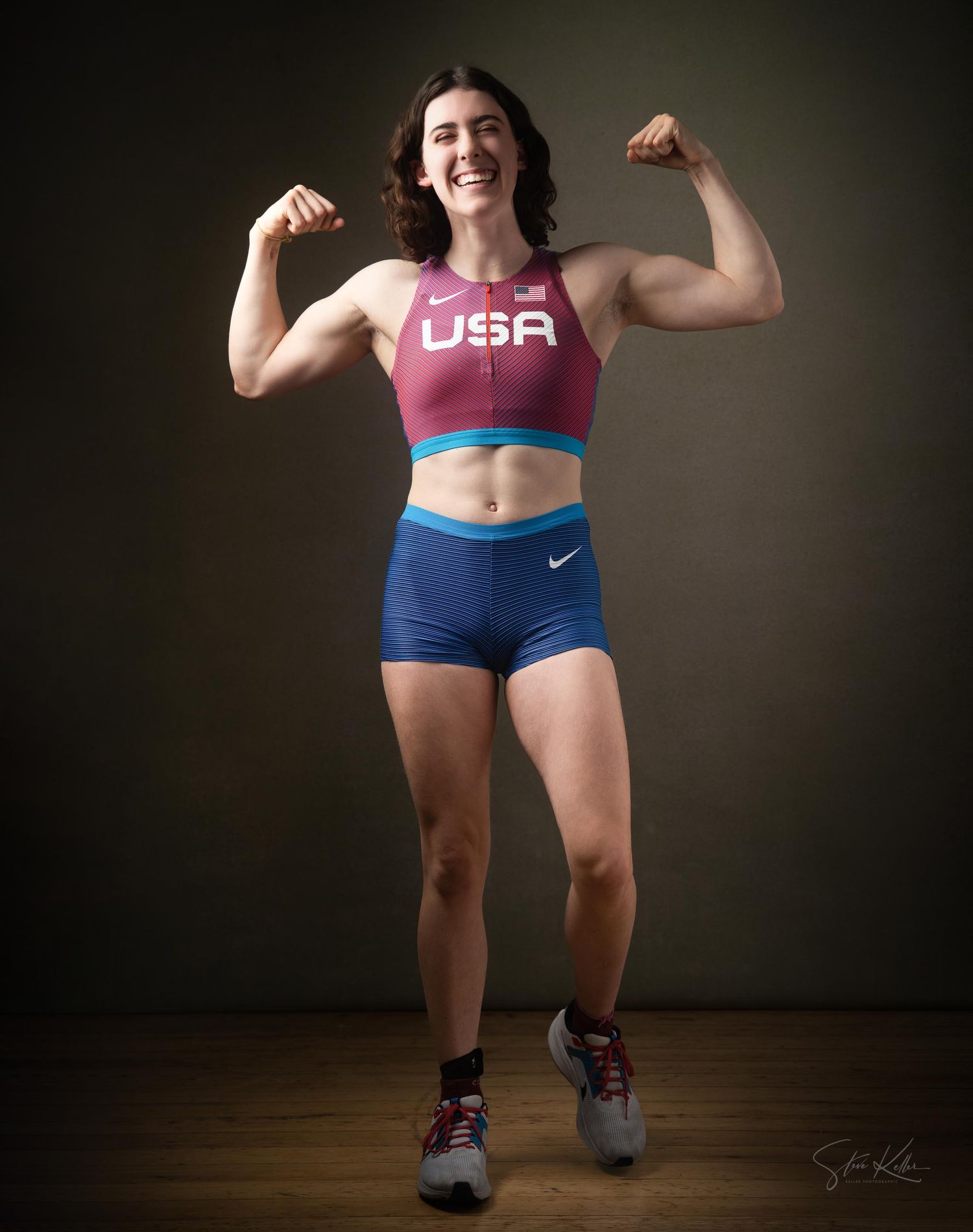 An athlete in USA team apparel strikes a strong flexing pose against a dark backdrop during a fitness photoshoot.