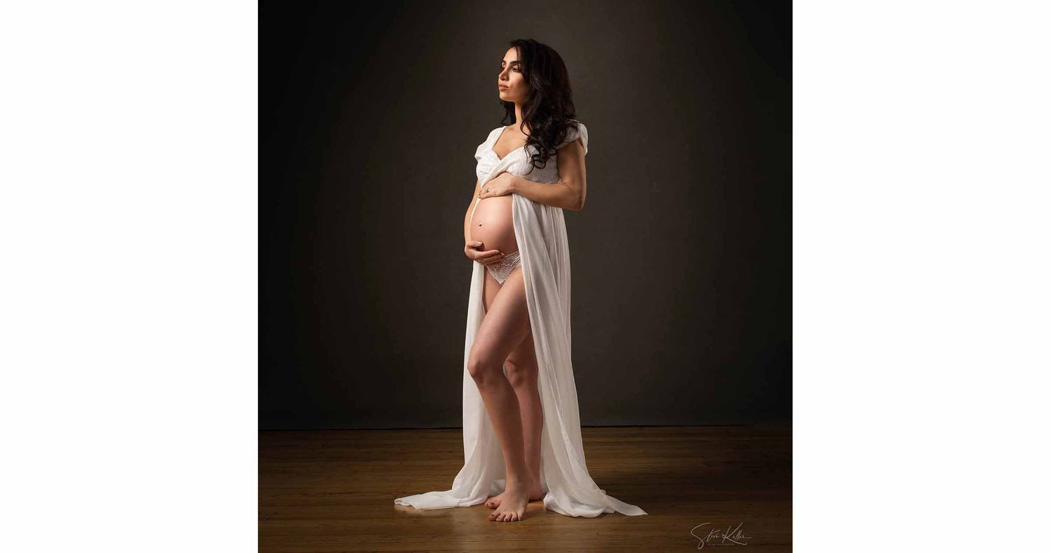 Pregnant woman with long dark hair stands in a studio draped in flowing white fabric, cradling her baby bump in a maternity portrait.