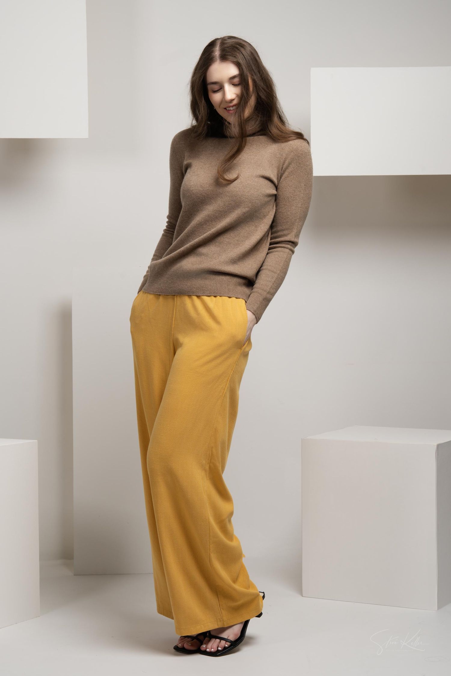 Woman in brown turtleneck sweater and yellow wide-leg pants posing against neutral studio backdrop.